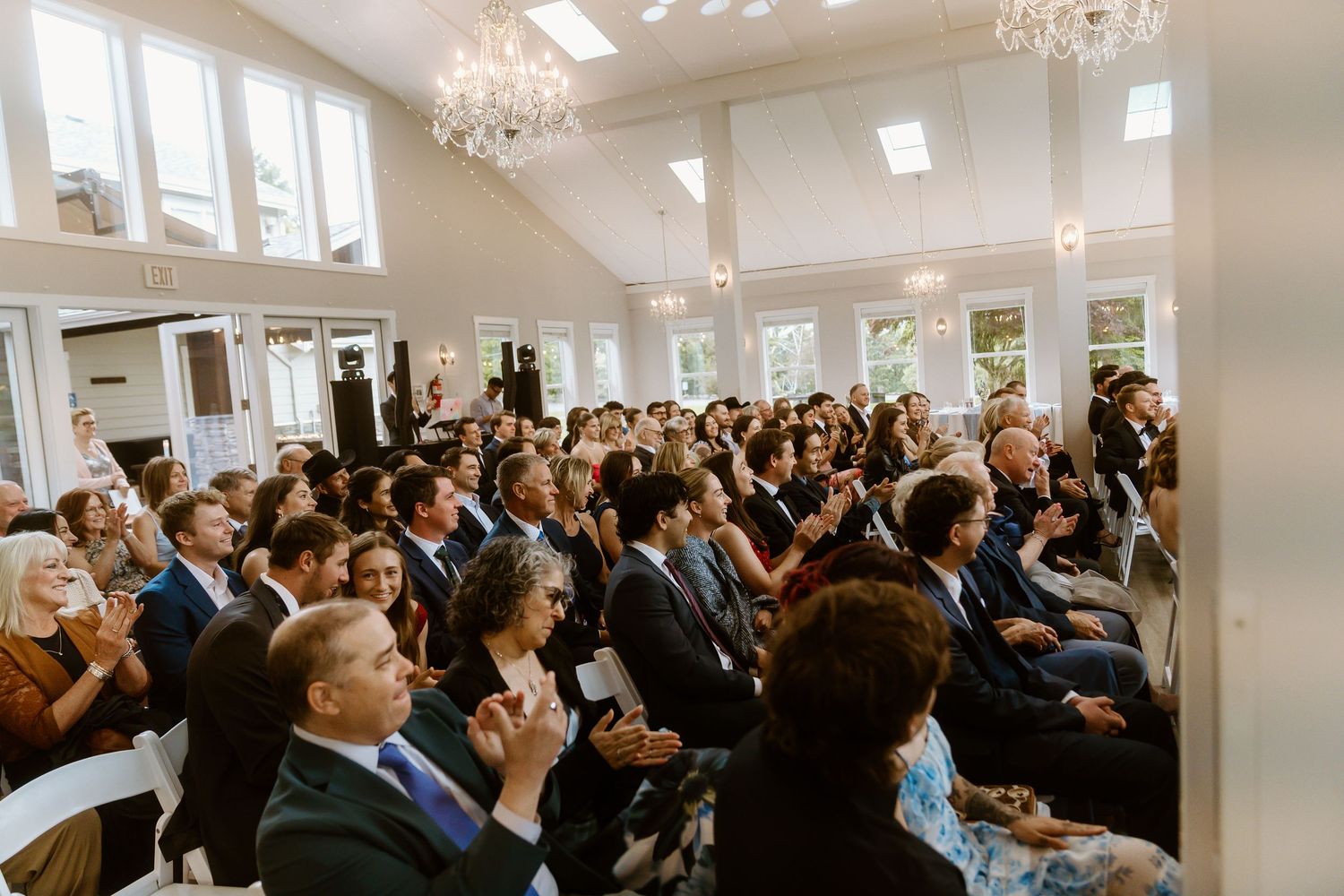 Guests seated in a bright white wedding venue with chandeliers, watching an indoor ceremony with large windows.