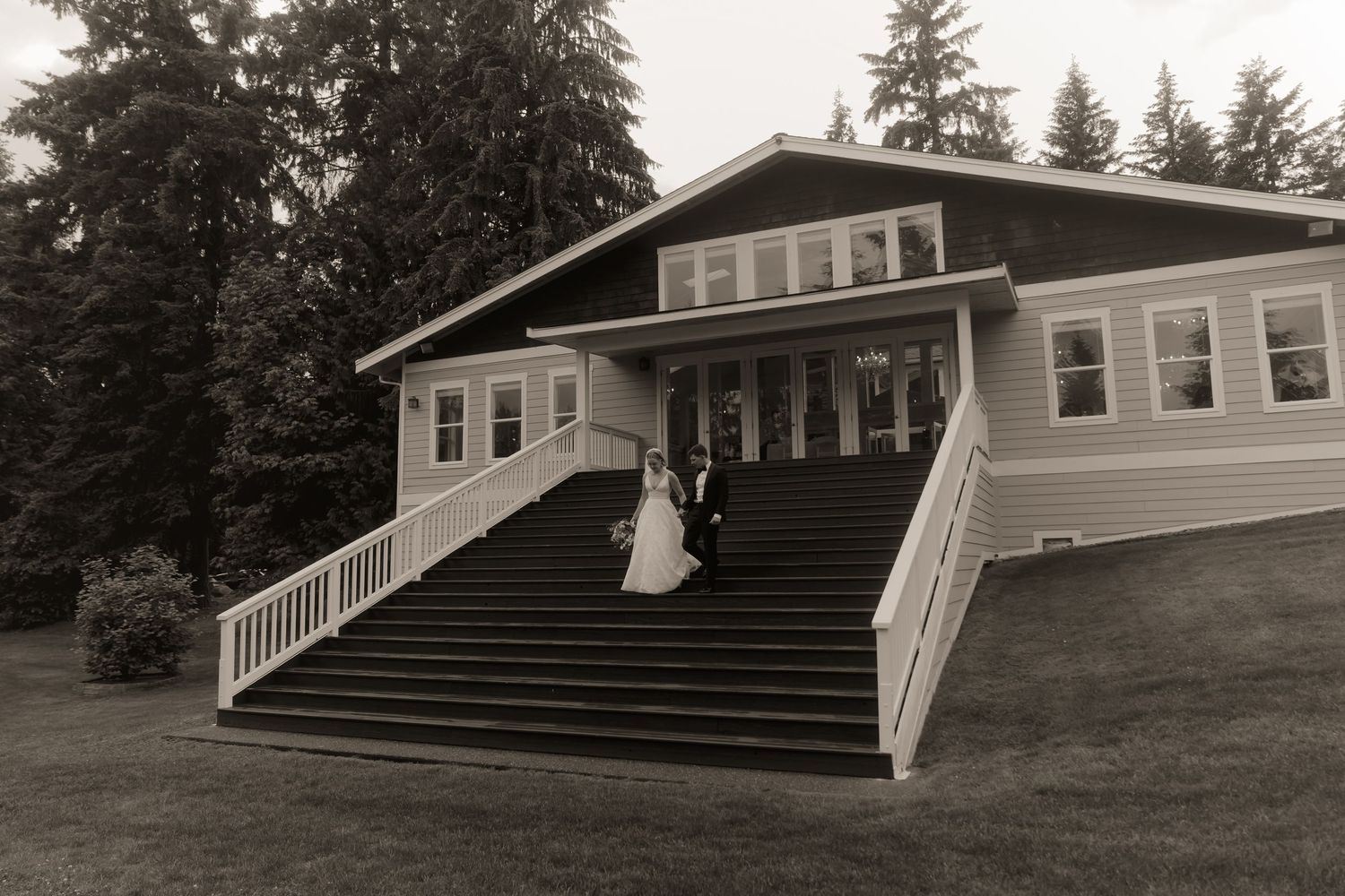 Black and white photo of a bride and groom on grand staircase outside elegant wedding venue surrounded by tall trees.