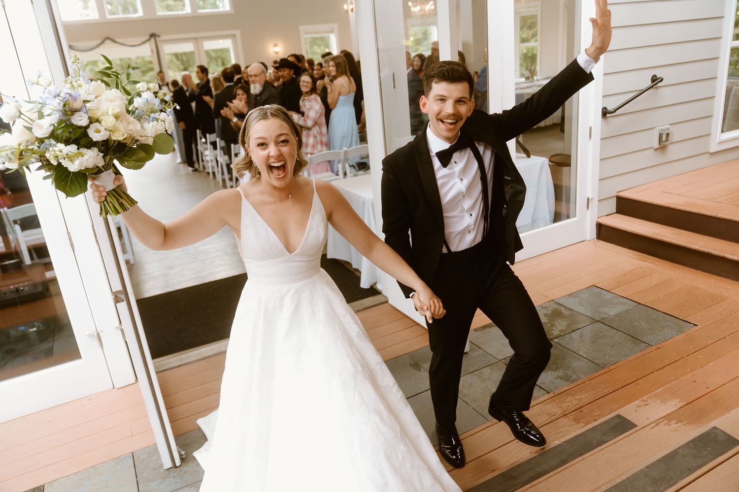 Joyful newlyweds celebrate after wedding ceremony, bride in white gown holding bouquet, groom in black tuxedo, guests in background.