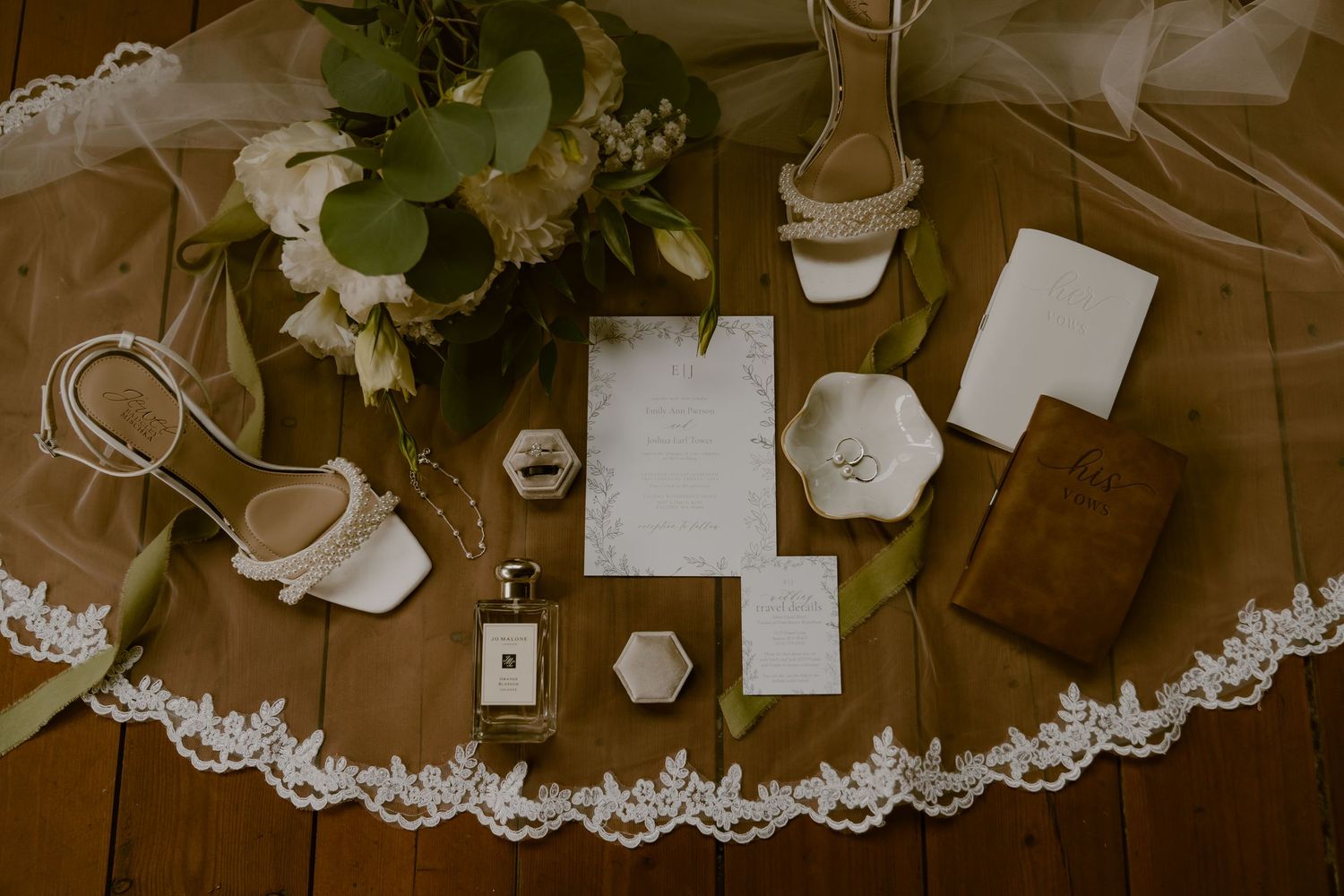 Wedding details flatlay with lace veil, stationery, shoes and floral arrangements on wooden surface.