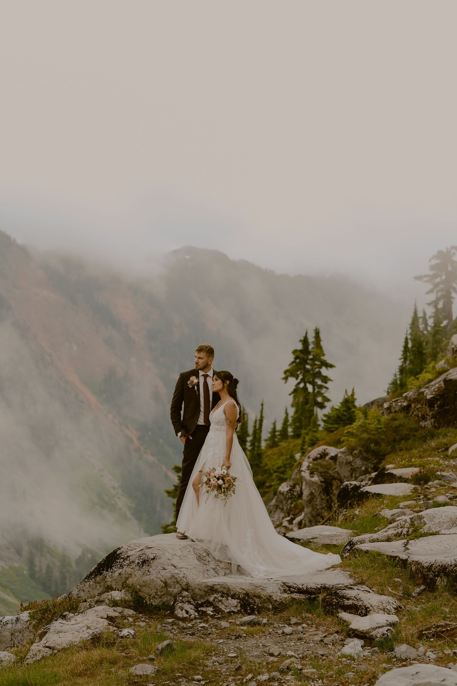 Wedding couple standing on rocky mountain overlook with dramatic foggy views.