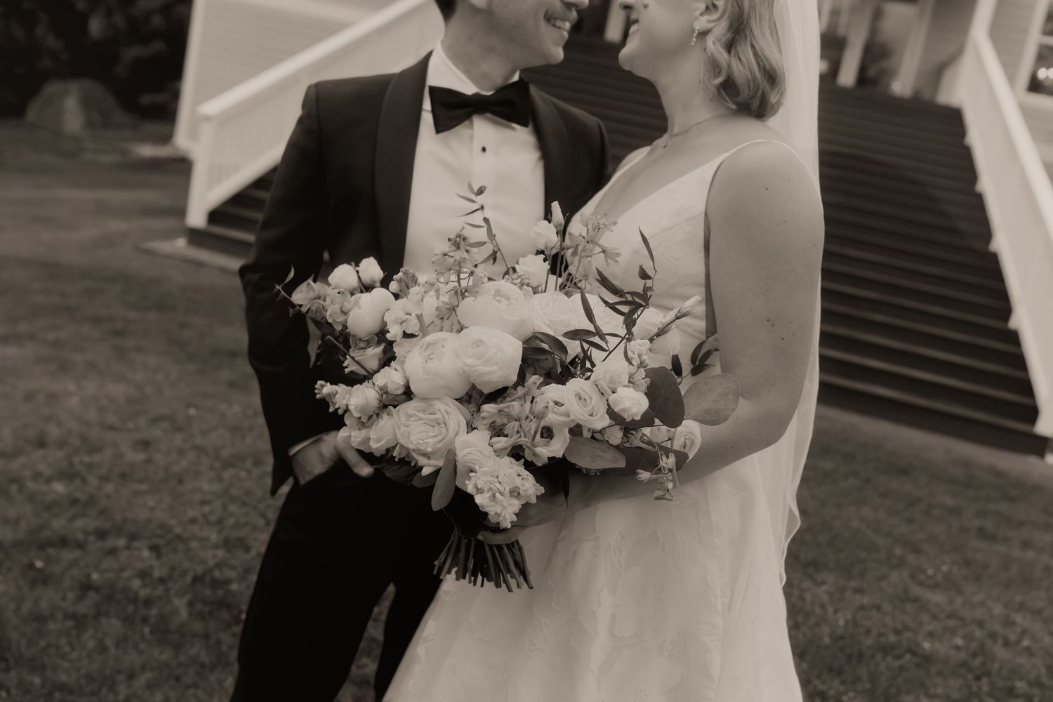 Black and white wedding photo of bride holding floral bouquet and groom in tuxedo standing together outdoors.