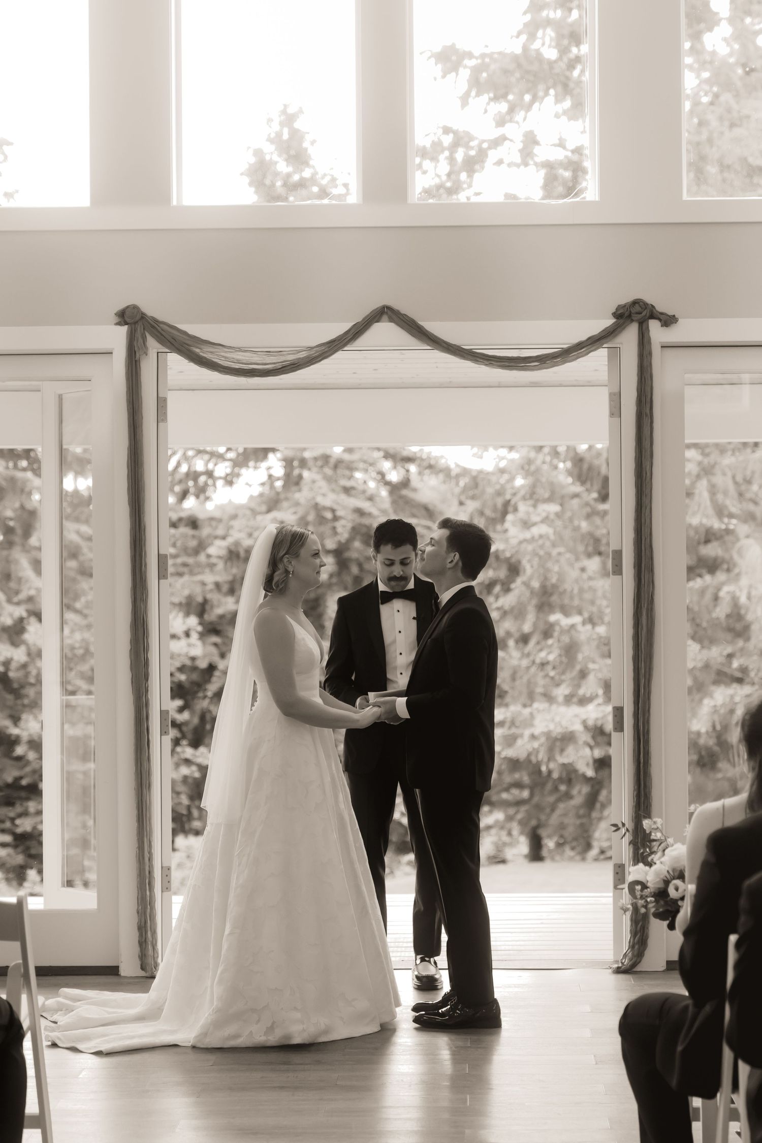 Black and white photo of bride and groom exchanging vows at indoor wedding ceremony with large windows.