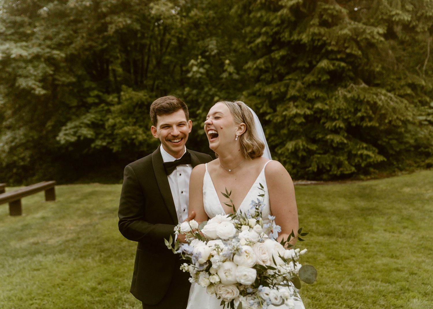 Laughing bride in white gown with veil and groom in black tuxedo holding bouquet outdoors.