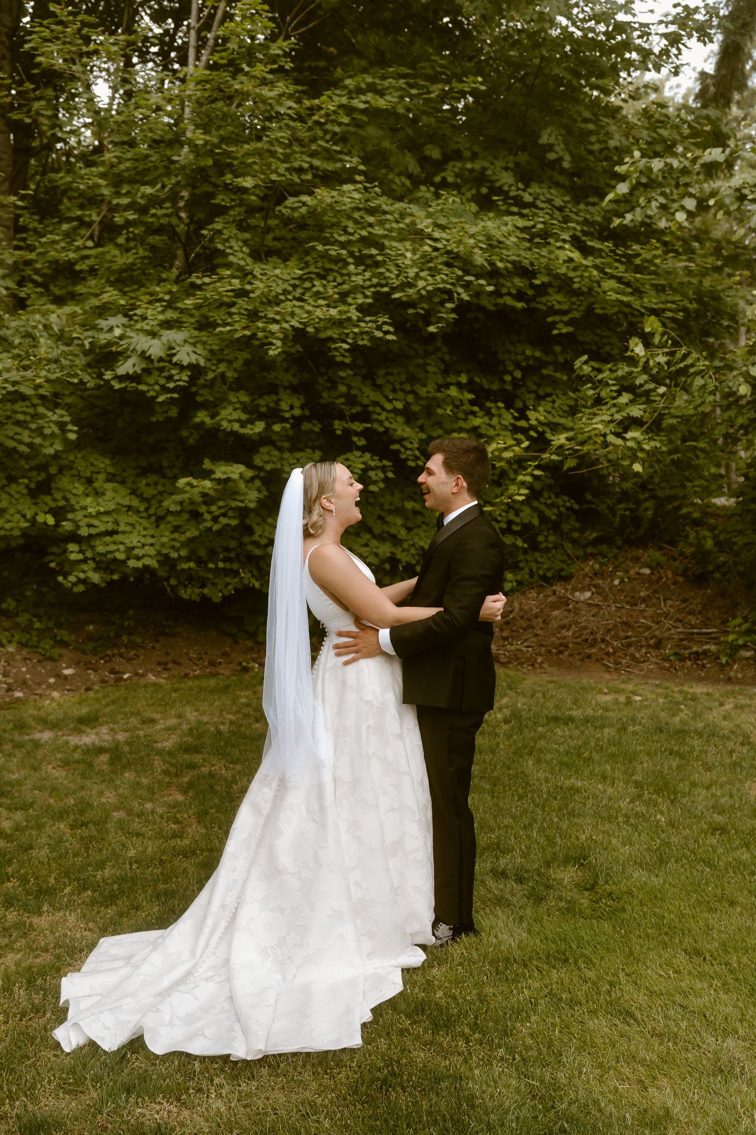 Bride in white lace gown with veil and groom in black suit share first look outdoors.