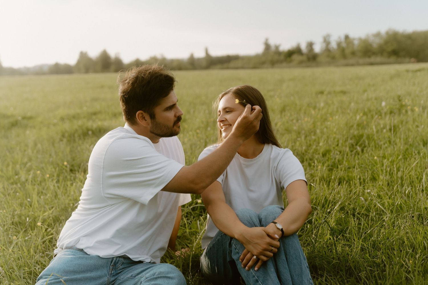 Couple sitting in green field, man gently touching woman's face in tender romantic moment outdoors.