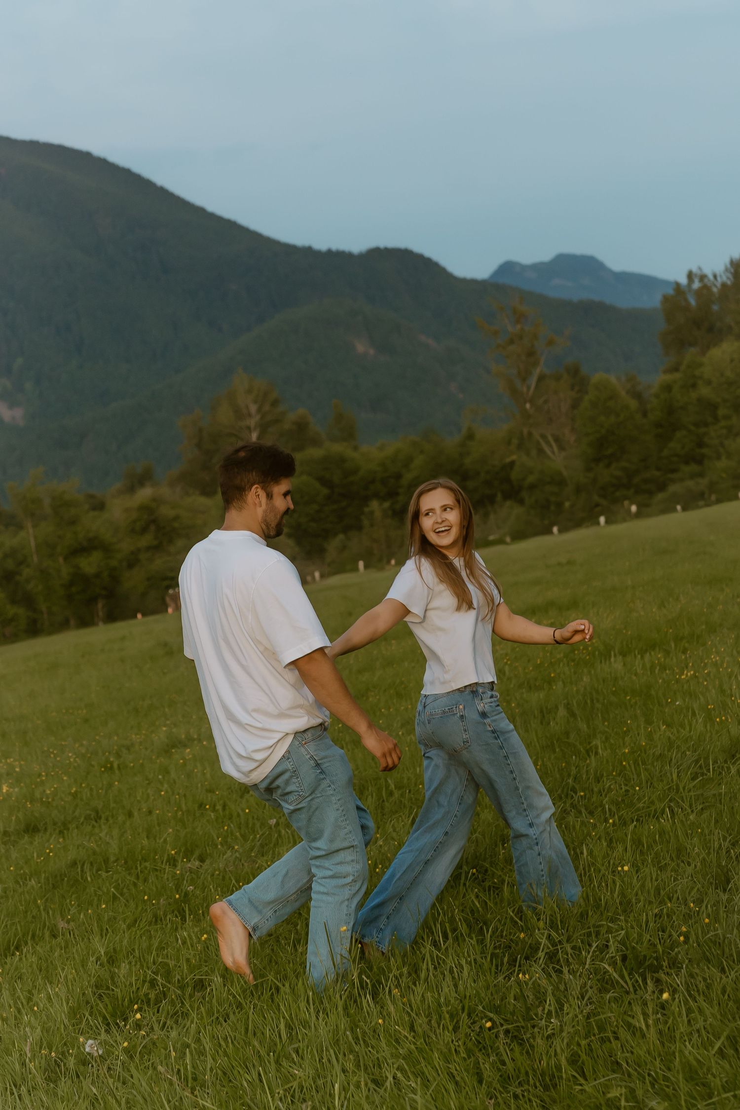Couple holding hands dancing in green mountain meadow, wearing matching white shirts and blue jeans.