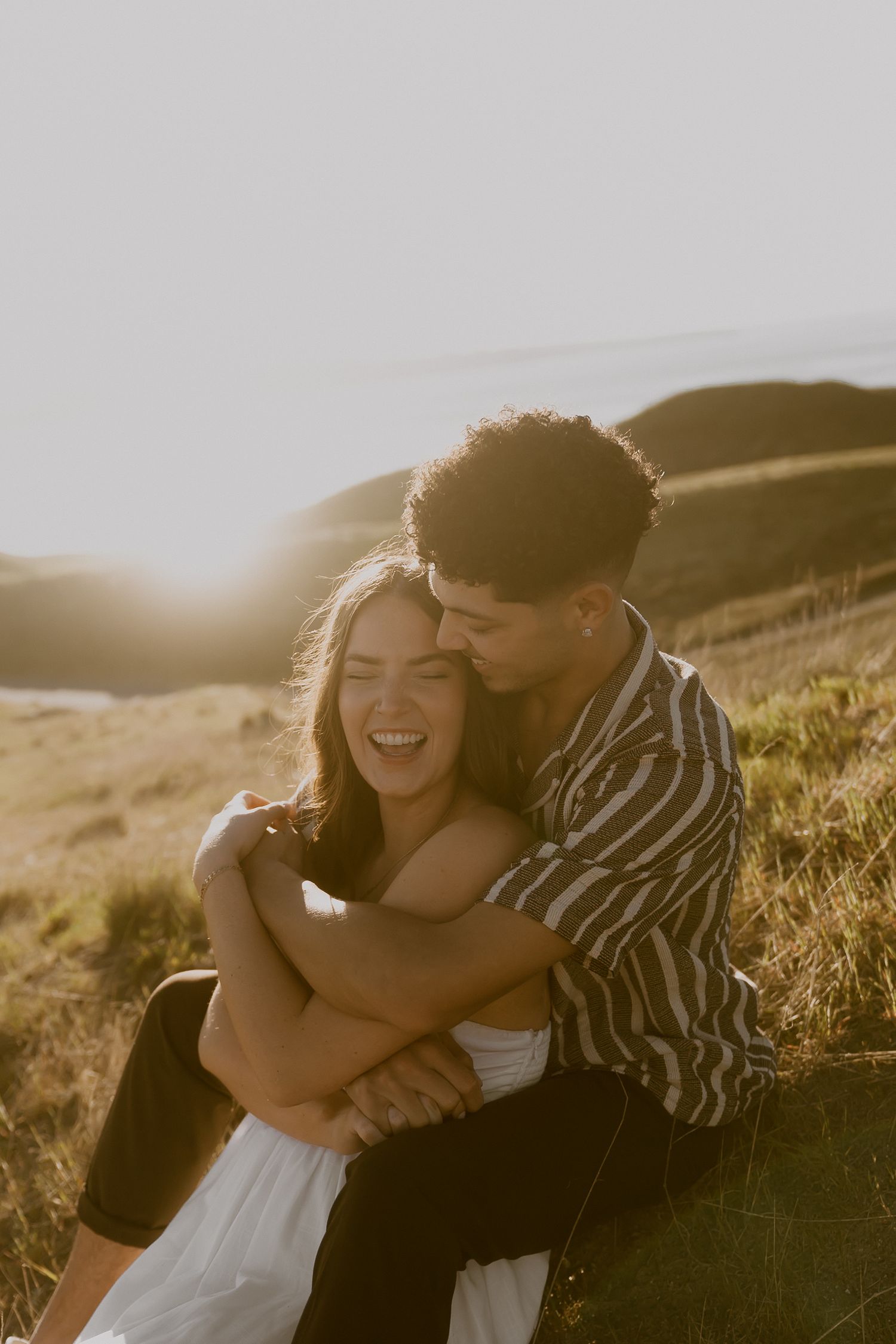 A laughing couple embraces at golden hour on a grassy hillside, the man hugging the woman from behind.