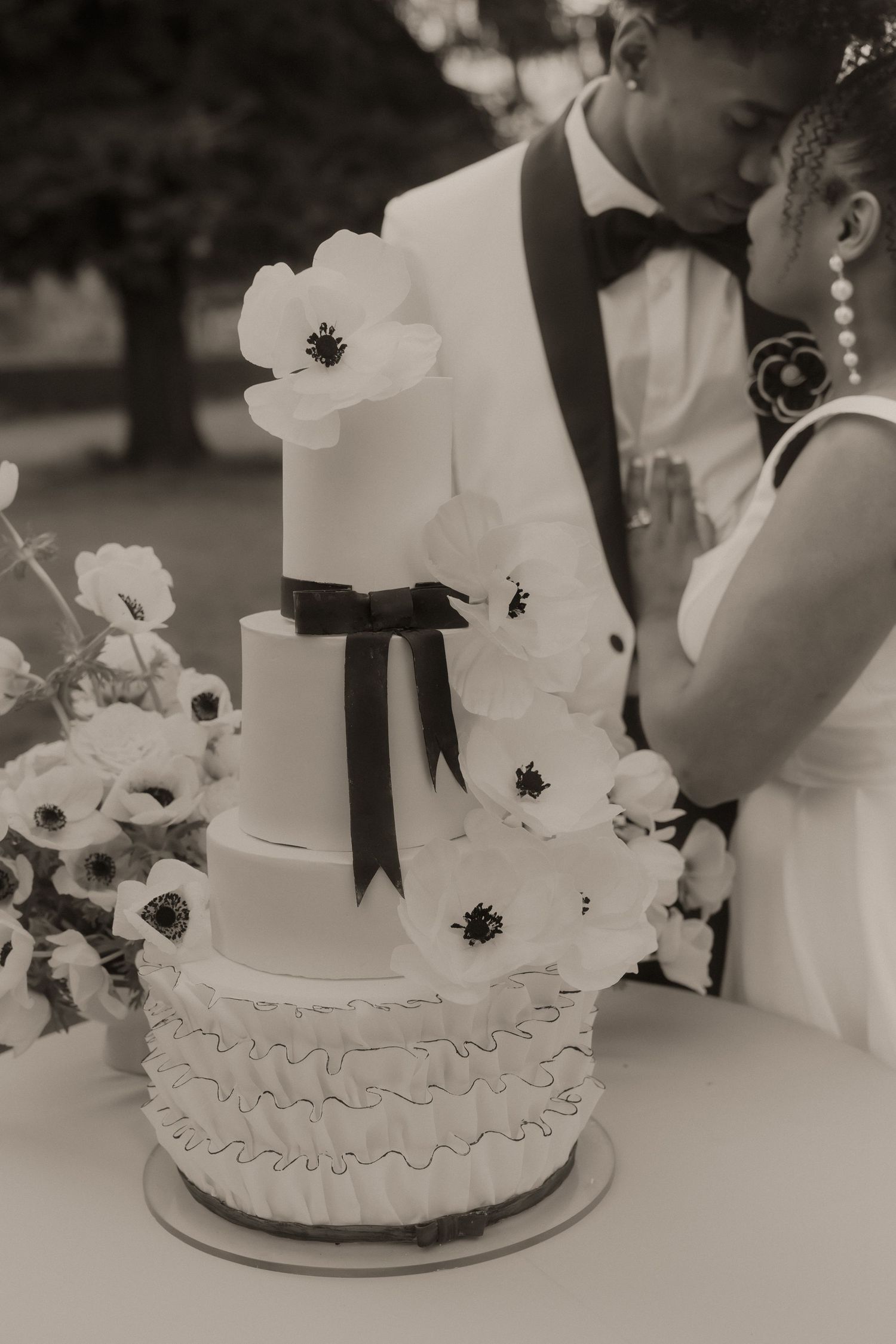 Elegant black and white photo of a couple cutting a tiered wedding cake adorned with anemone flowers and a black ribbon.