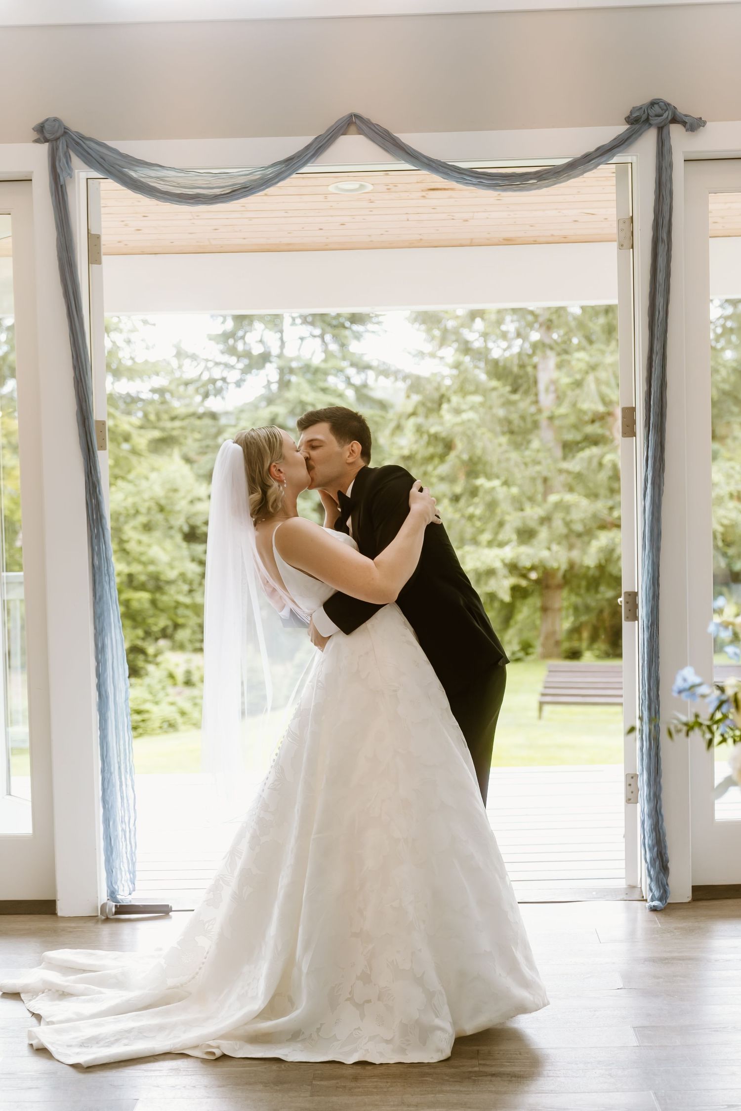 Bride and groom share first kiss under blue draped doorway in bright elegant wedding venue.