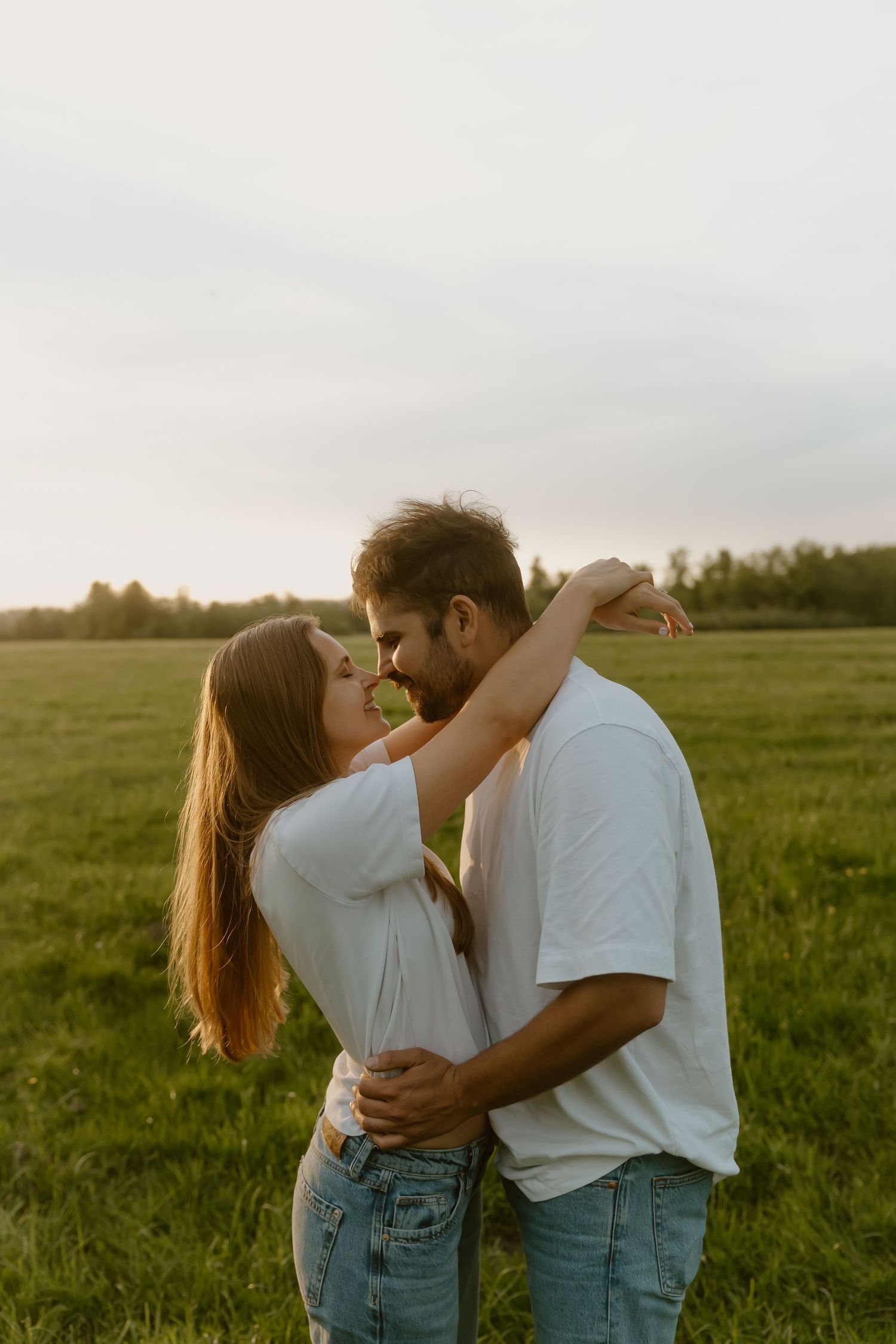 Couple embracing in a green field at golden hour, wearing white tops and jeans, about to kiss.