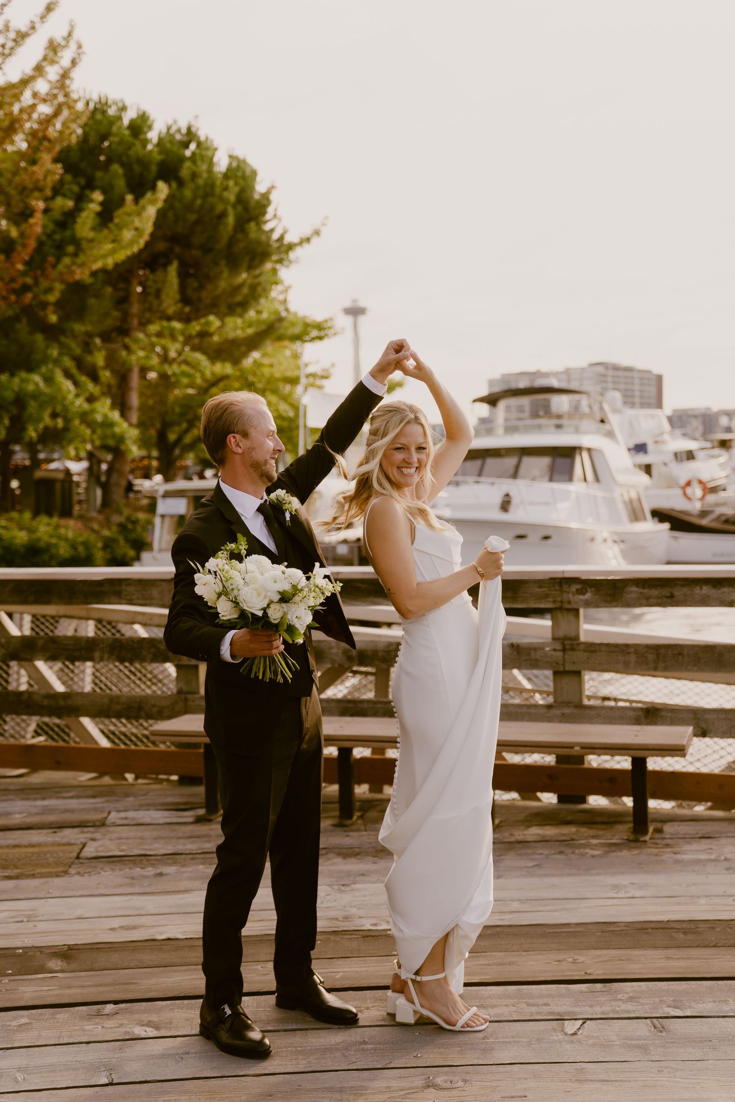 A couple dancing and twirling together on a wooden dock by a marina at sunset.