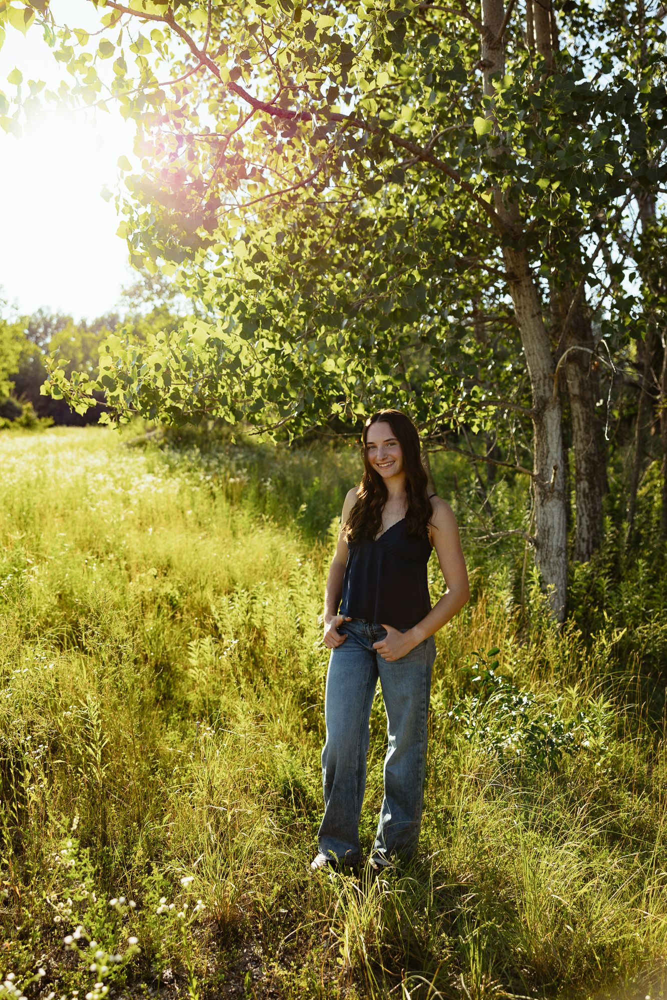 Young woman in black top and jeans smiling outdoors near birch tree in sunlit green meadow.