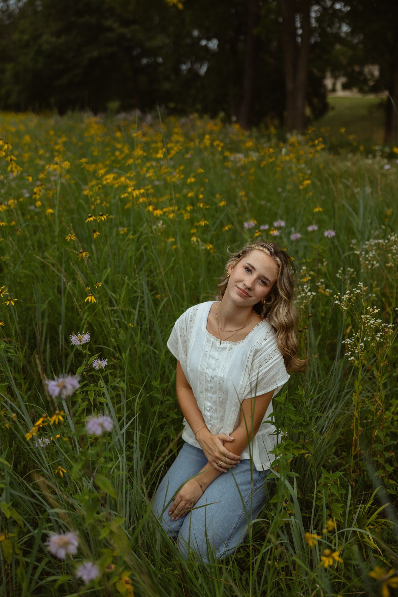 A person kneels in a wildflower meadow wearing a white blouse and jeans surrounded by yellow and purple blooms.