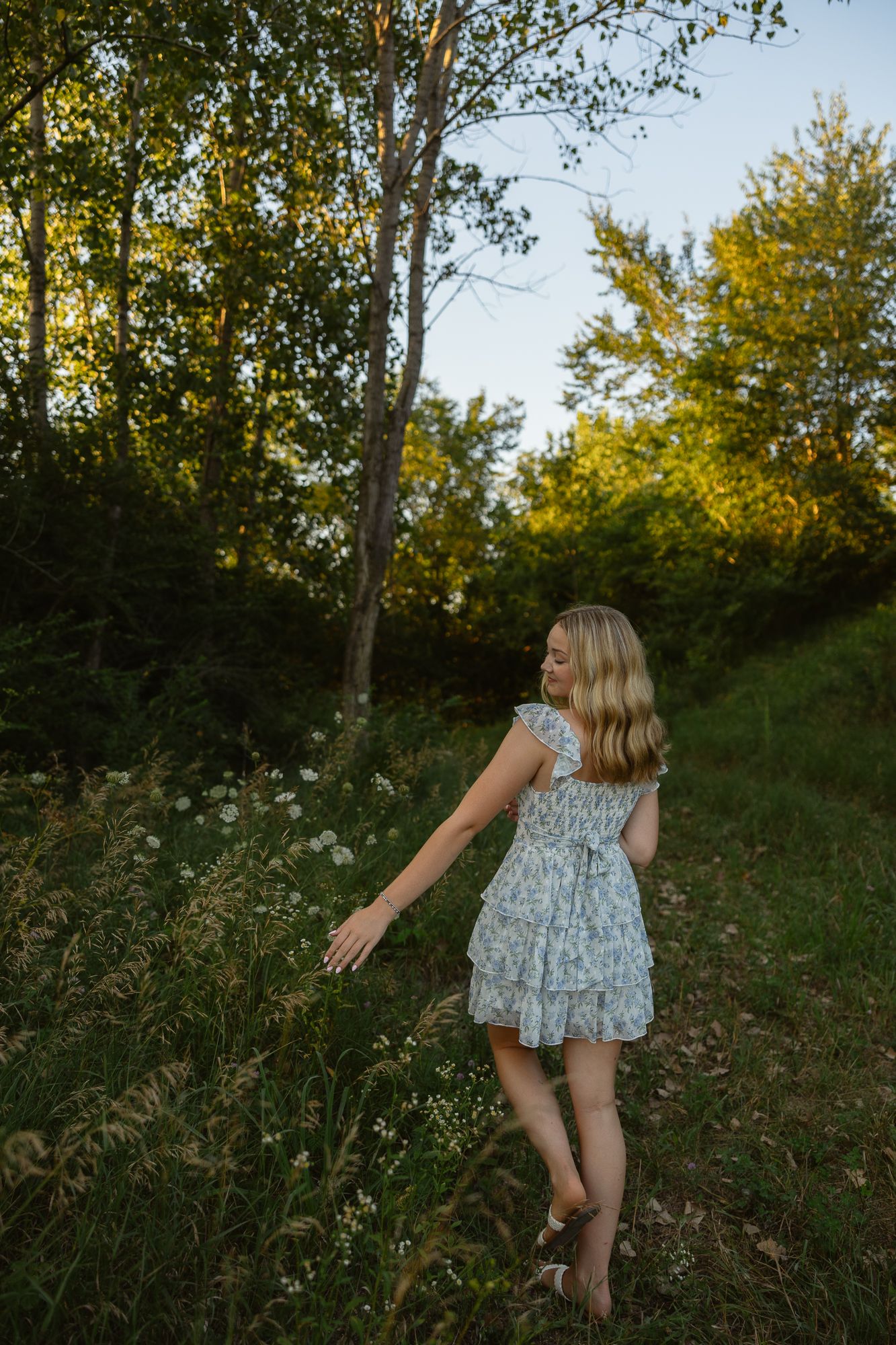 Young blonde girl in floral dress walking through wildflowers at golden hour in a lush green forest.