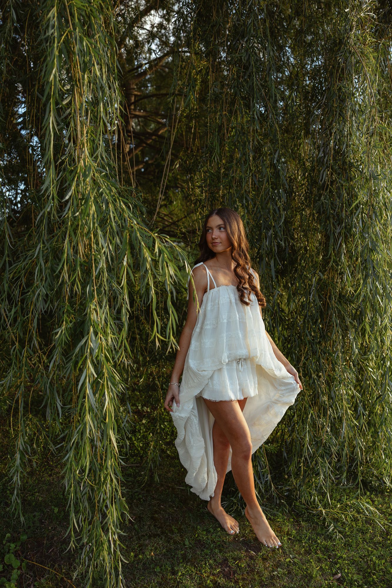 Young woman in white flowing dress standing barefoot among weeping willow branches in a lush green garden.