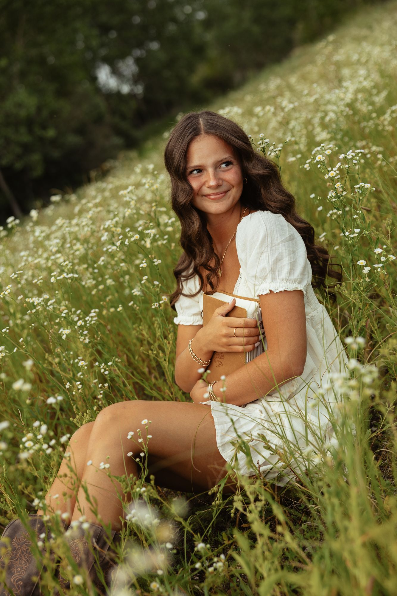 Young woman in white dress sitting in wildflower meadow, smiling and holding a book outdoors.