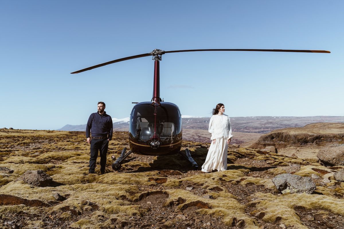 Couple standing beside a helicopter near a remote mountain lake in Hvalfjörður looking in opposite directions
