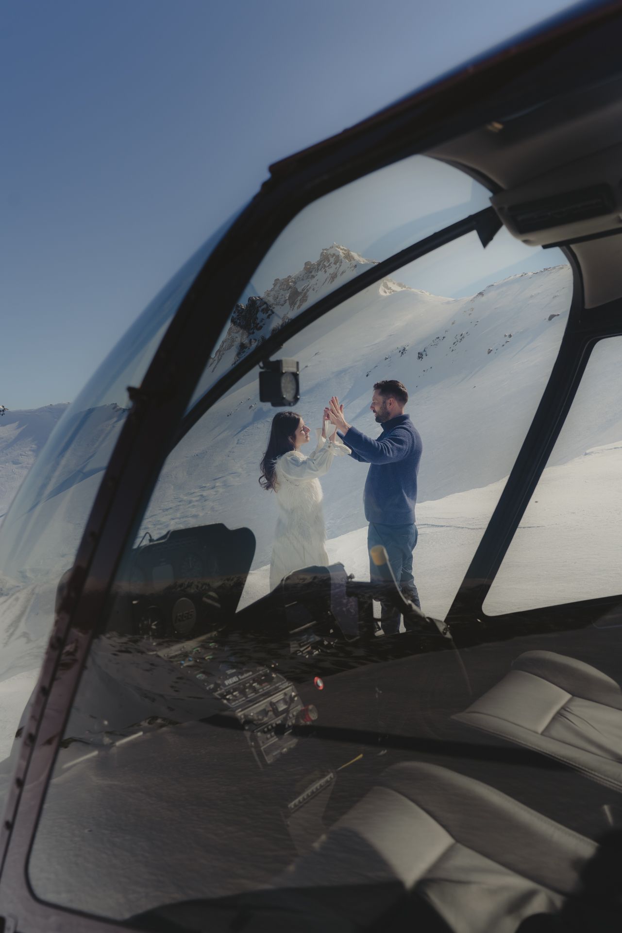 Couple dancing on a snowy mountain peak during a helicopter elopement seen through the helicopter window