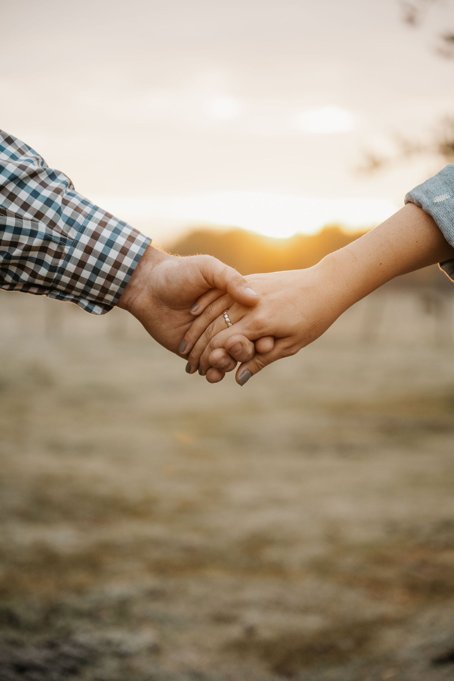 Two people holding hands at sunset with one person wearing a checkered shirt against a blurred outdoor background.