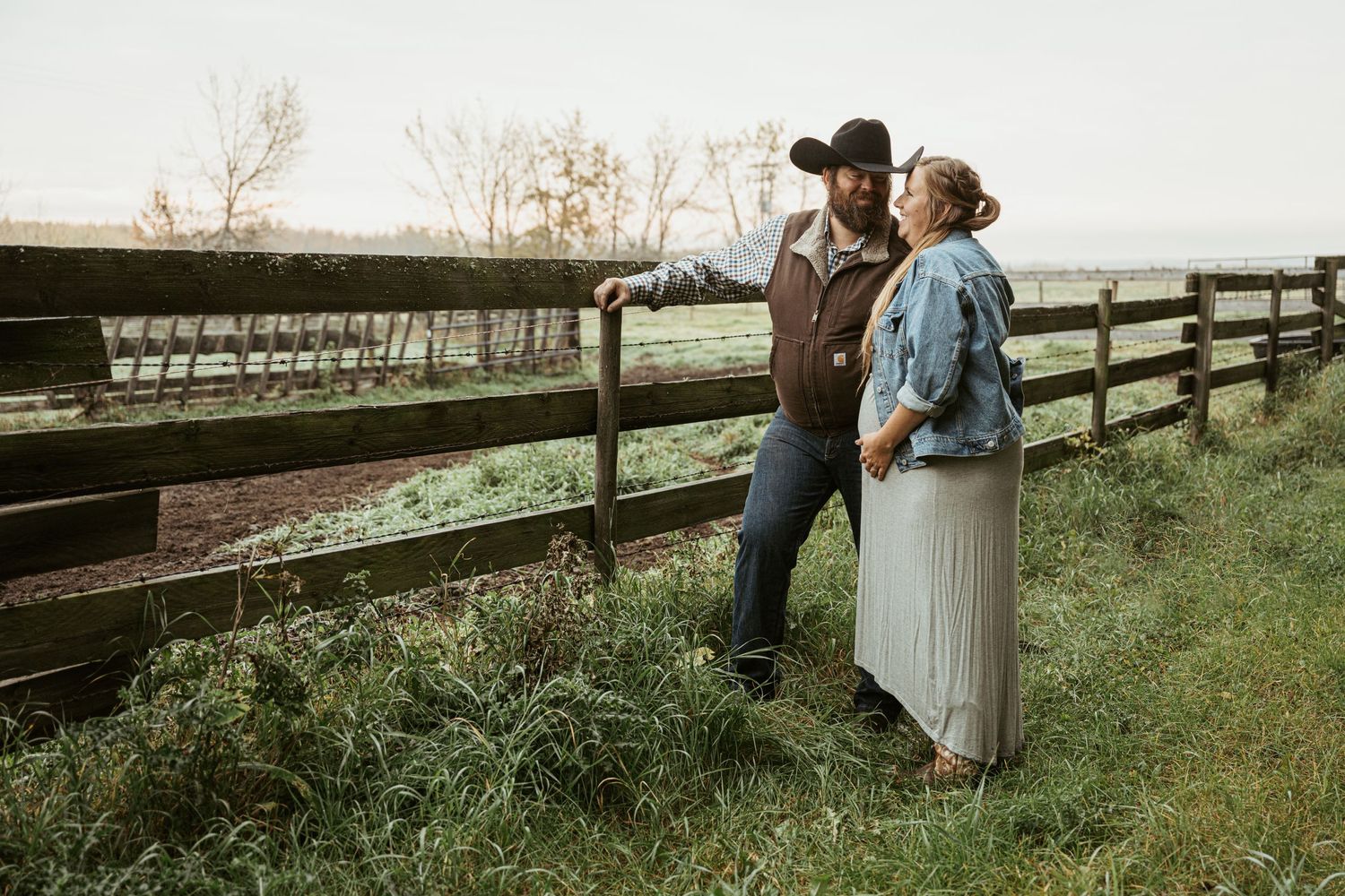 Two people in western attire lean against a wooden fence at sunset overlooking a countryside pasture.