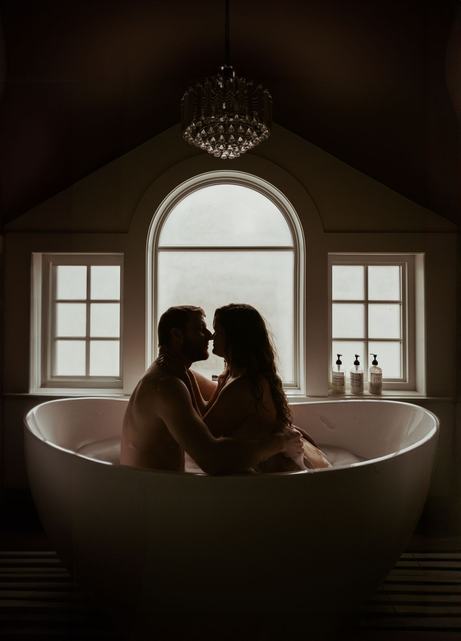 Silhouettes of a romantic couple share an intimate moment in a freestanding bathtub against an arched window backdrop.