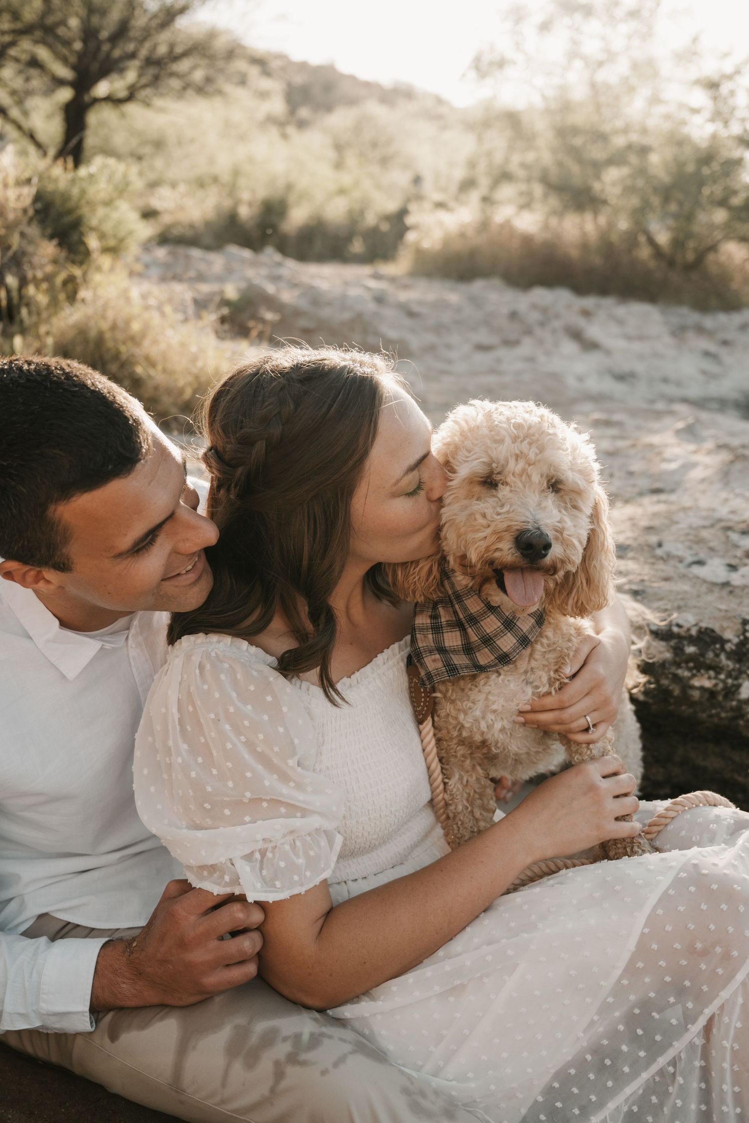 A couple shares a tender moment with their adorable Goldendoodle puppy wearing a plaid bandana in warm sunset lighting.