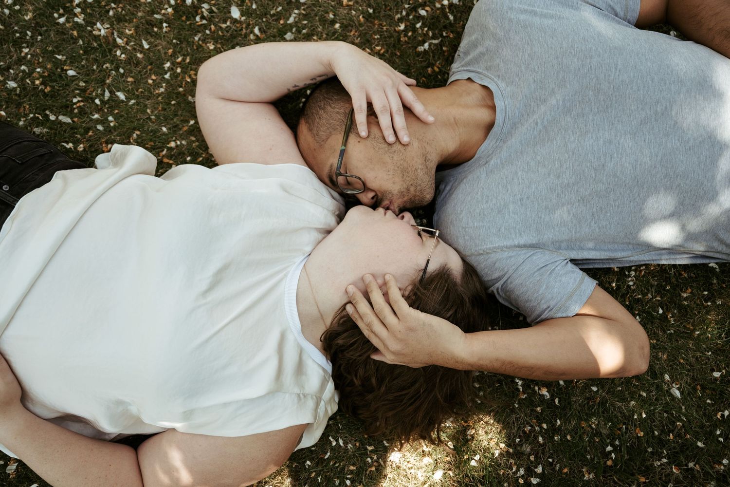 Two people lying on grass together in white and grey shirts, shot from above with dappled sunlight.
