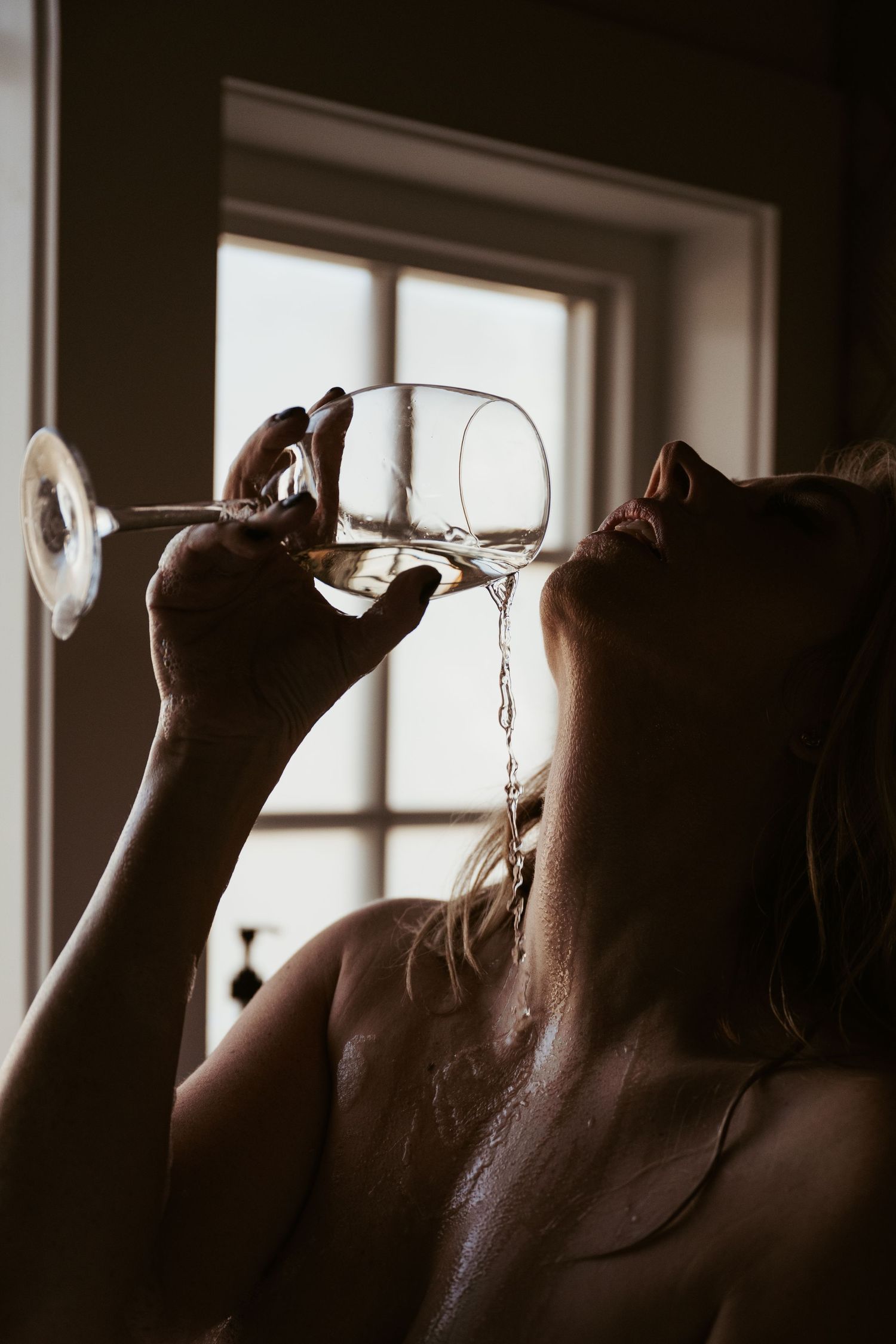 Artistic silhouette of someone pouring water from a wine glass in front of a sunlit window.