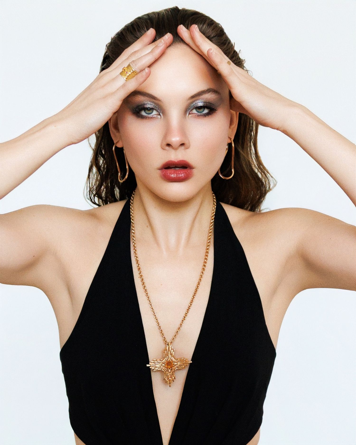 Fashion photo of model in black halter top wearing dramatic makeup and gold jewelry with hands posed on forehead.