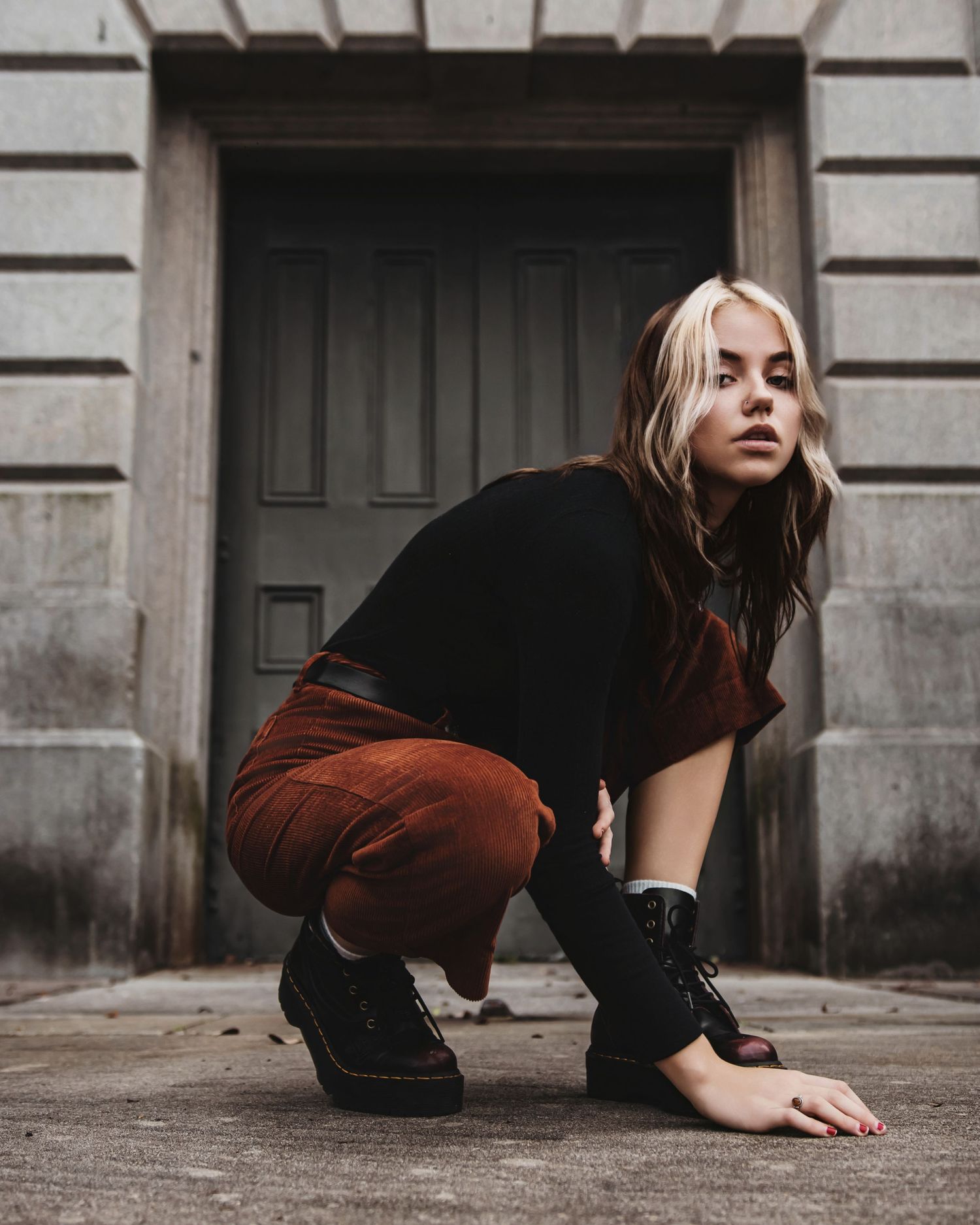 Artistic portrait of someone crouching in front of dark doorway wearing black top and rust-colored pants.