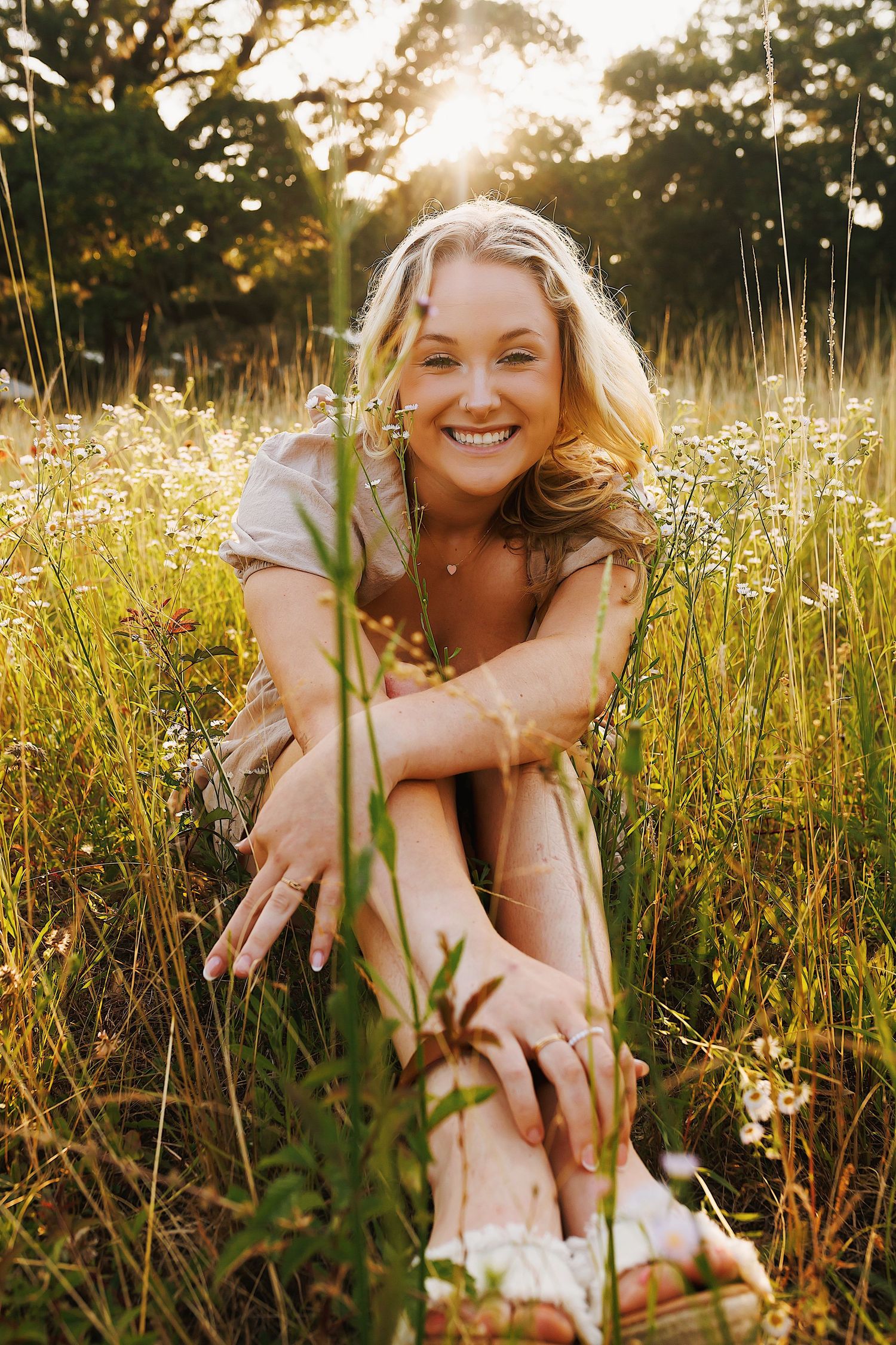 Person sitting in a golden sunlit meadow with tall grass and wildflowers during sunset.