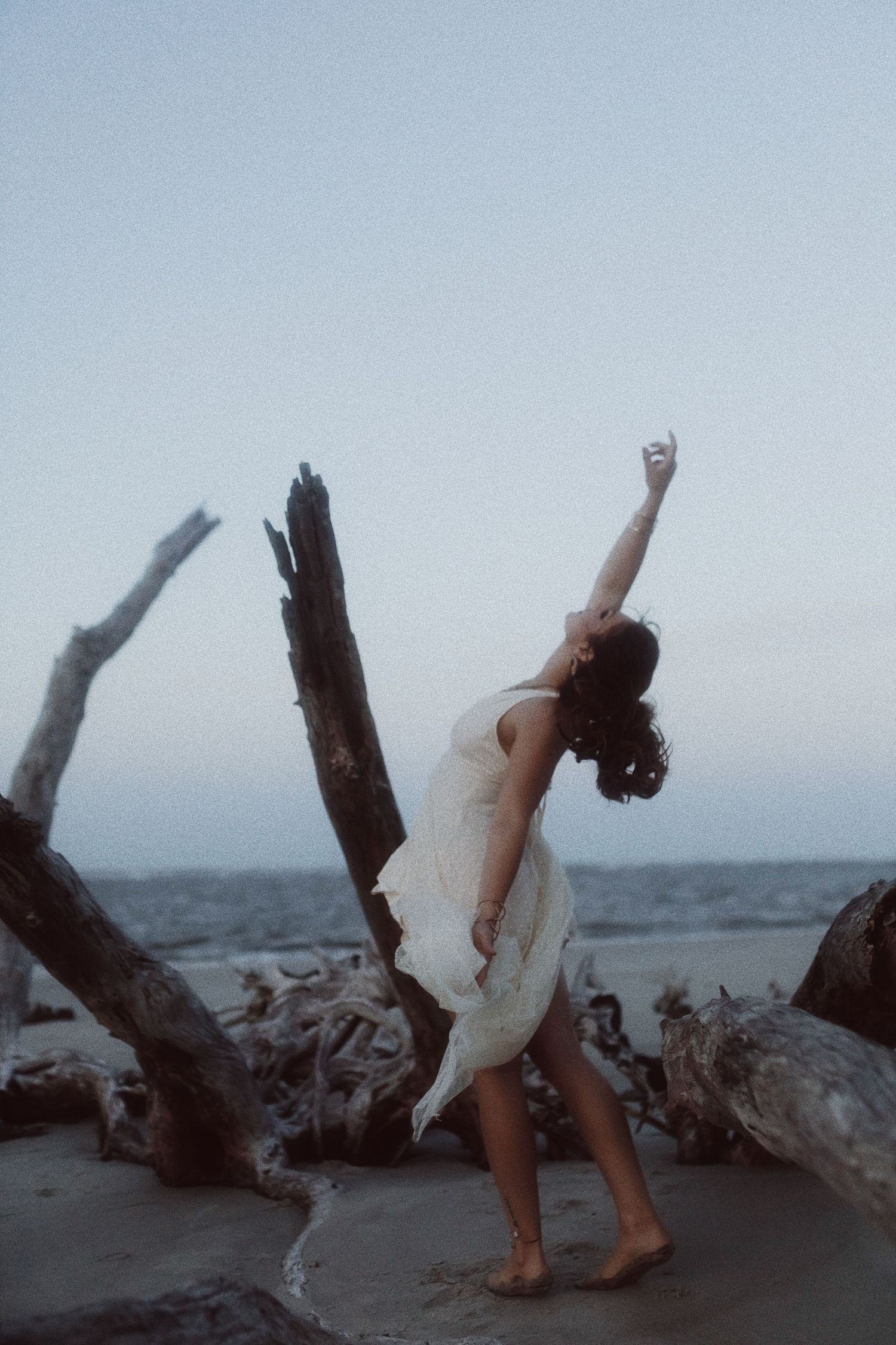 A dancer in a flowing white dress performs an elegant backbend pose on a beach with driftwood at dusk.