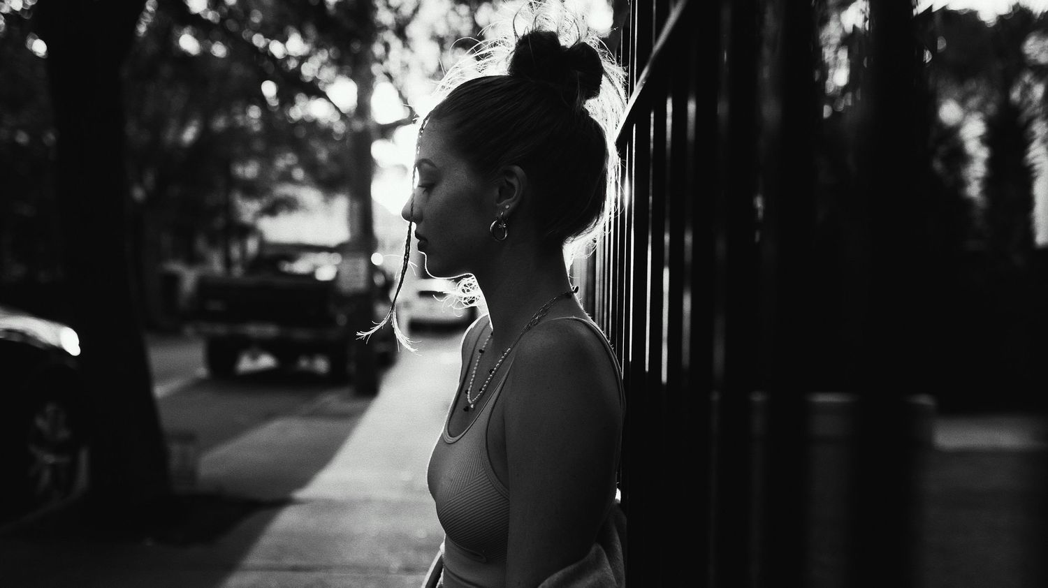 Black and white silhouette of a person in profile standing outdoors during sunset with trees and buildings visible.