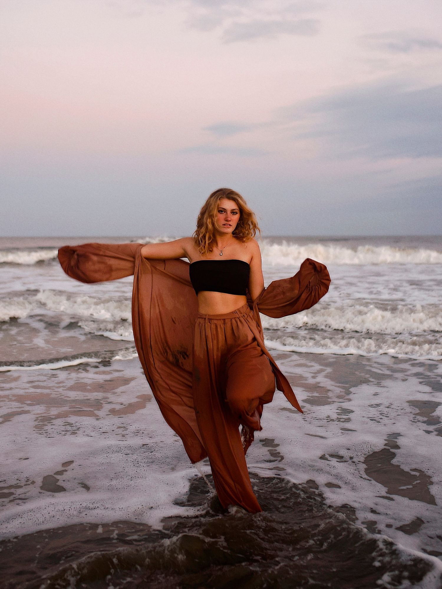 A figure in a flowing brown dress poses gracefully at the ocean's edge as waves crash against the shore at sunset.