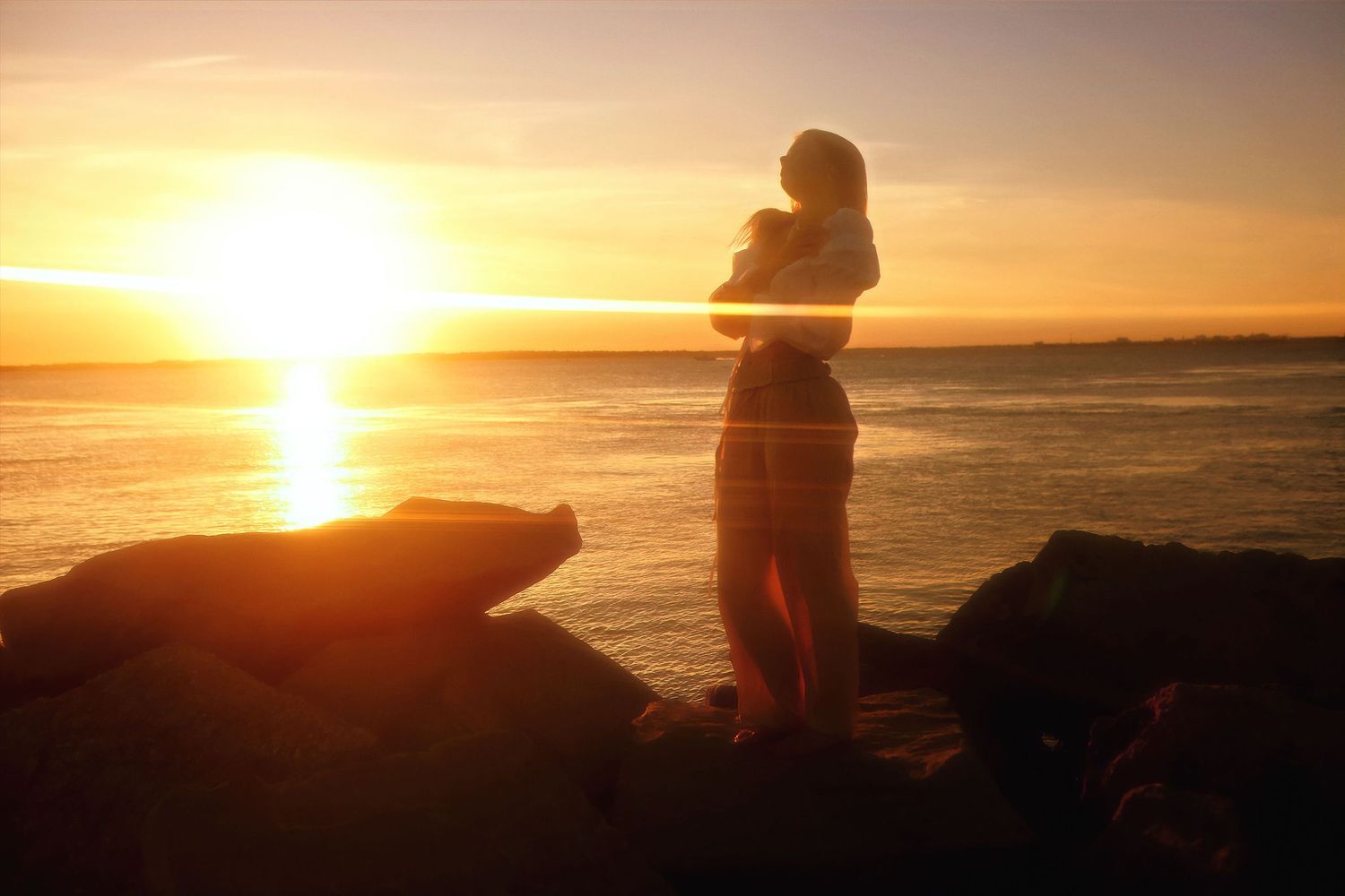 Silhouetted figures watching a golden sunset over a tranquil ocean from a rocky coastal viewpoint.