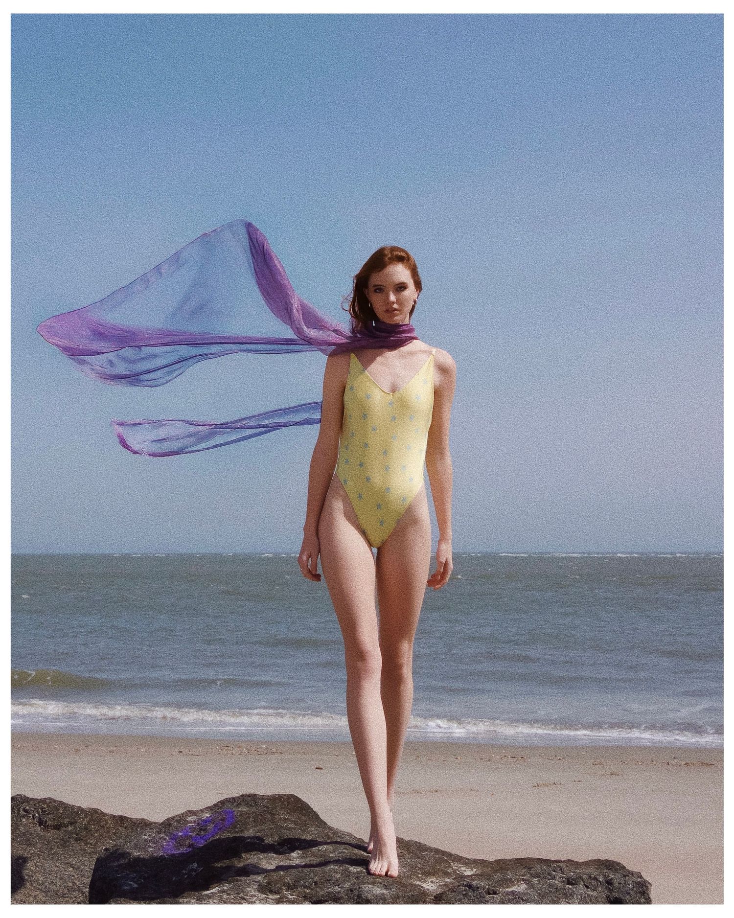 A figure in a yellow swimsuit poses on a beach while a purple scarf billows in the wind.