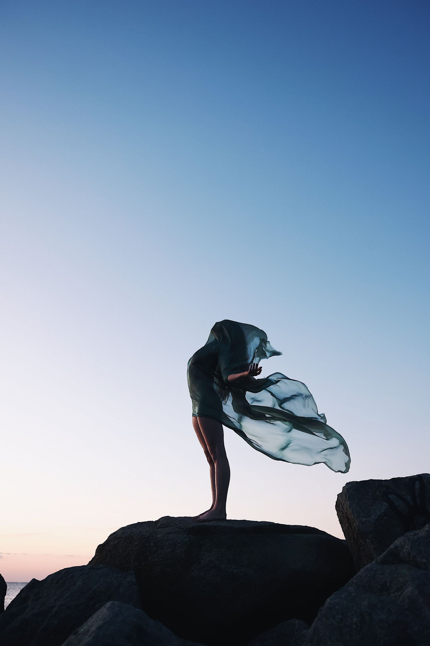 Silhouette of a person standing on rocks at sunset with flowing fabric billowing in the wind against blue sky.