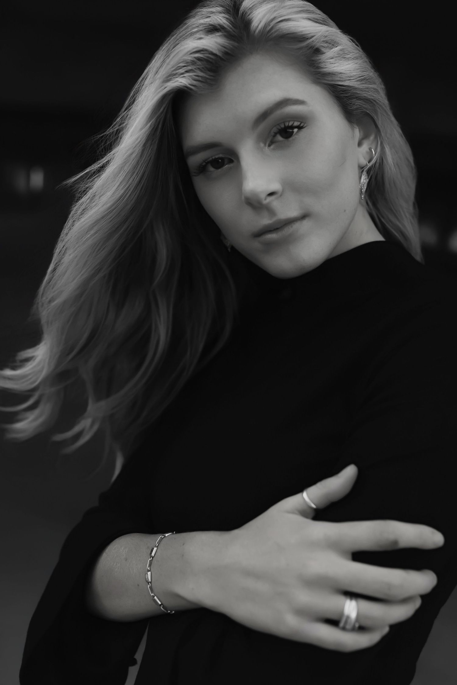Black and white portrait of a person wearing a black turtleneck sweater and delicate jewelry against a dark background.