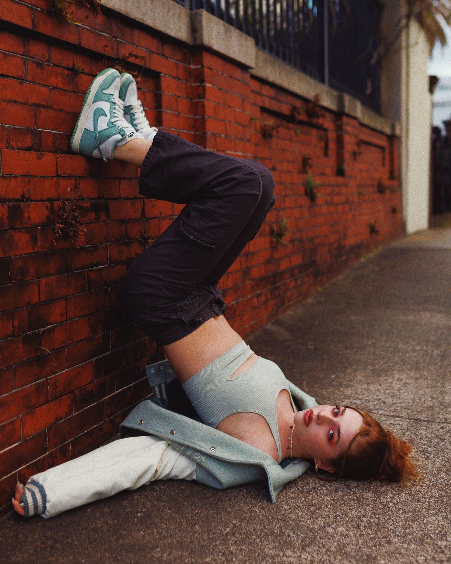 Person in casual attire lies sideways against red brick wall, one leg propped up displaying teal and white sneakers.