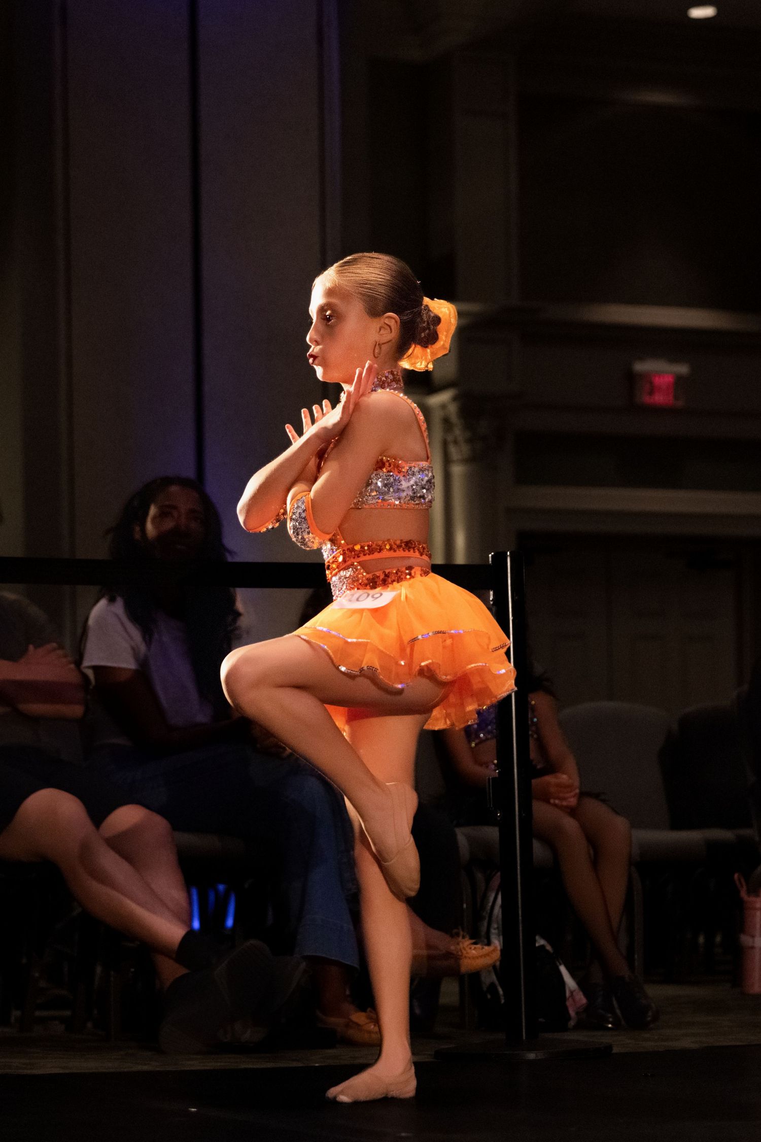 A dancer in a bright orange costume performs an energetic pose during a stage performance in a dimly lit venue.