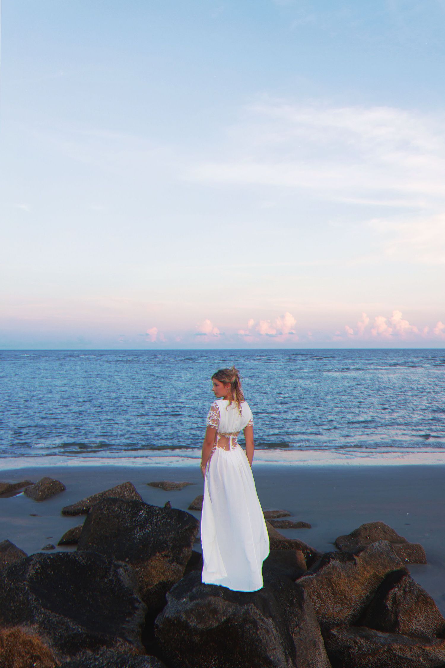 Scenic ocean view at sunset with dark rocks along the beach shore and soft pastel colors in the sky.