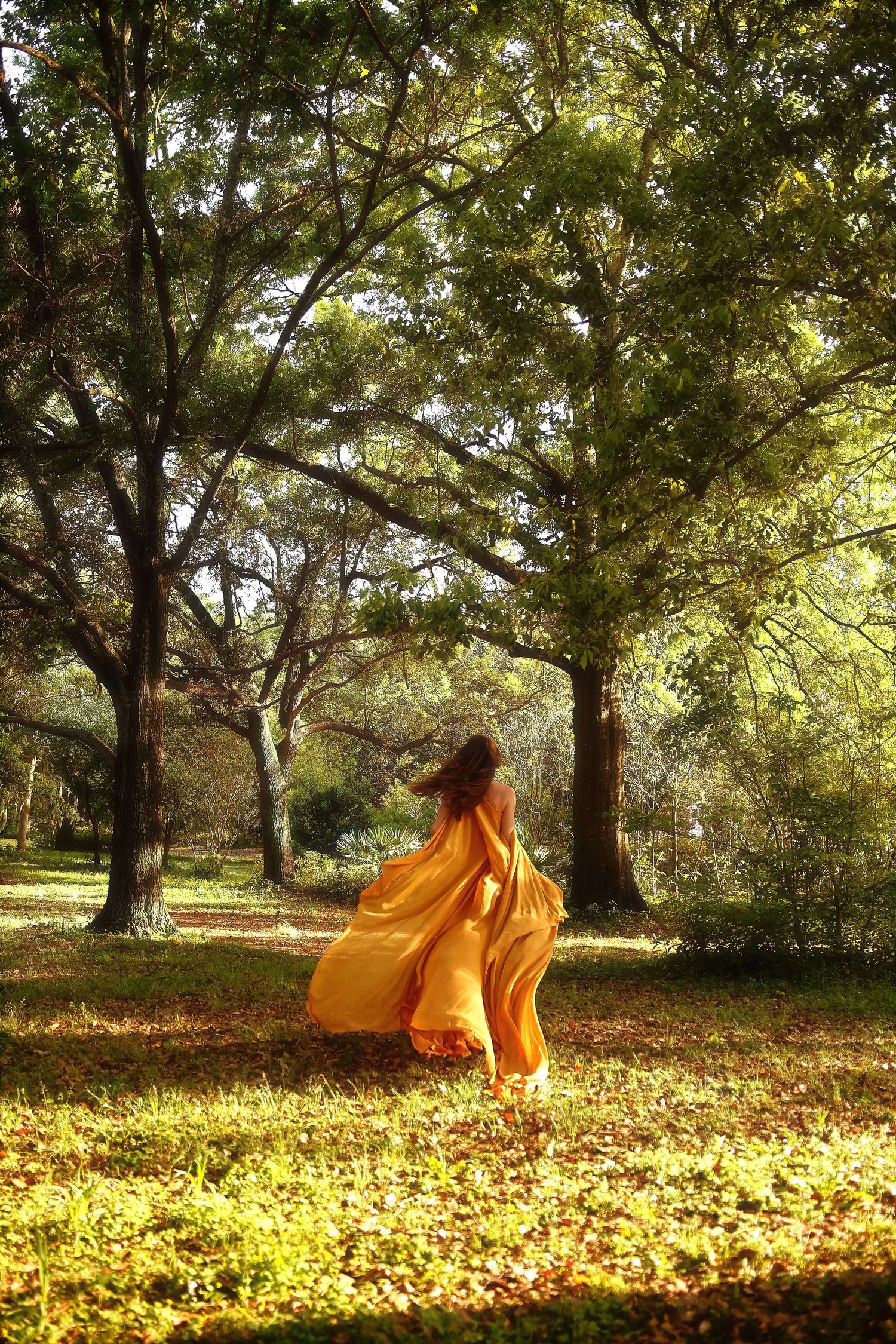 A figure in a flowing orange dress twirls gracefully among autumn trees with fallen leaves scattered on the ground.