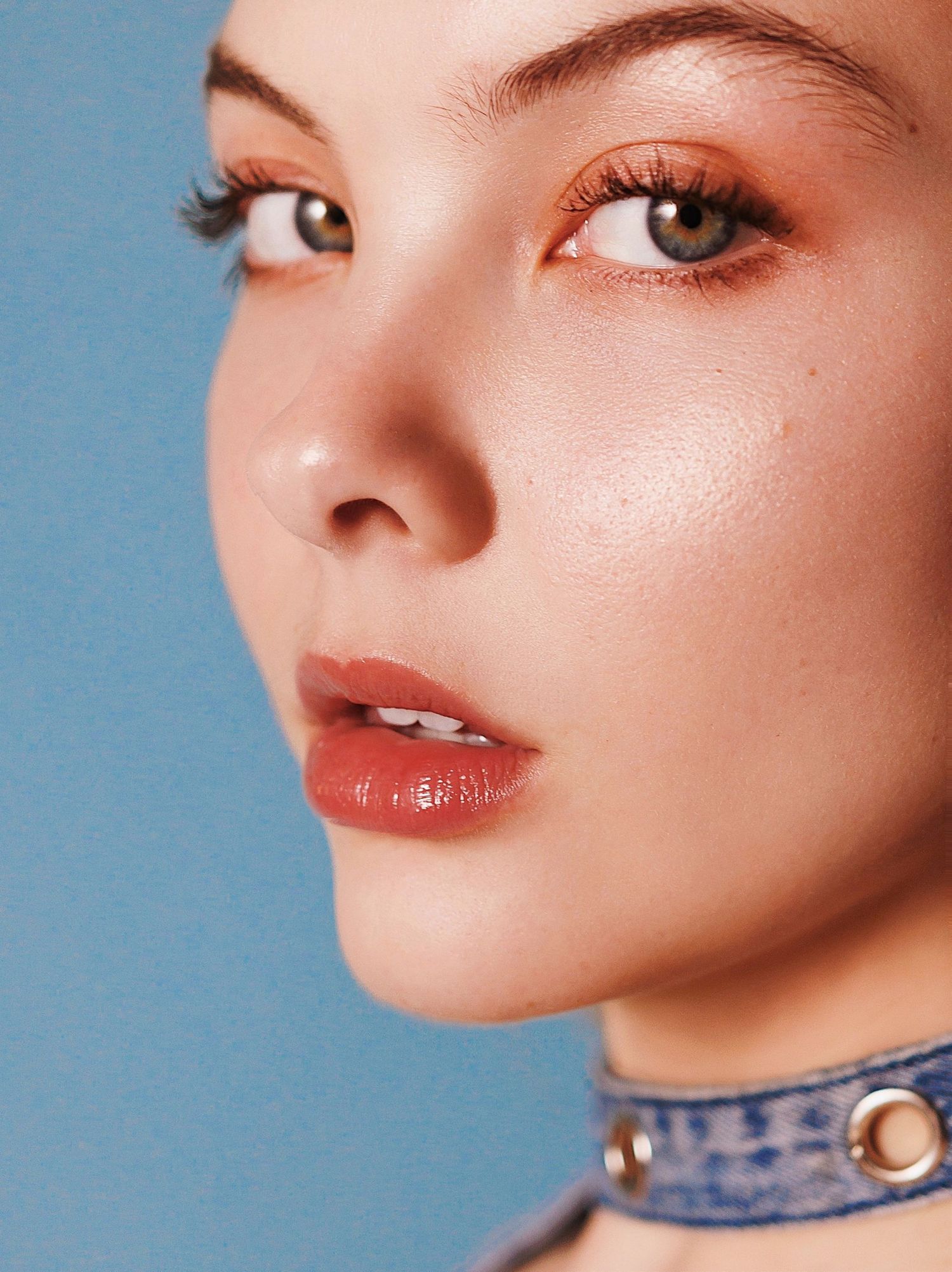 Close up beauty portrait with clear glowing skin, rose gold eyeshadow and coral lips against blue background.