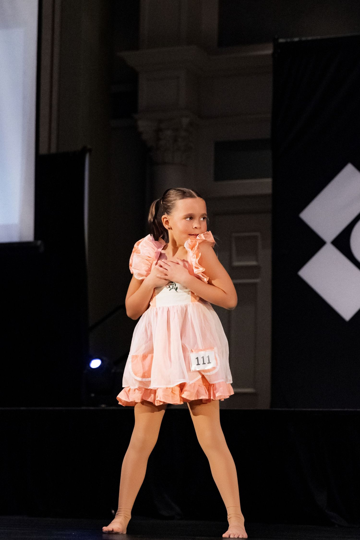 Young performer in a peach-colored dress strikes an expressive dance pose on a dark stage during a competition.