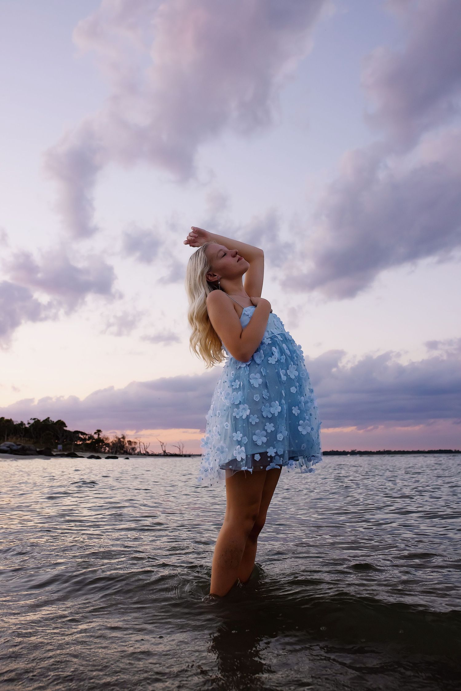 A person in a blue dress stands in calm ocean waters at sunset while silhouetted against a cloudy sky.
