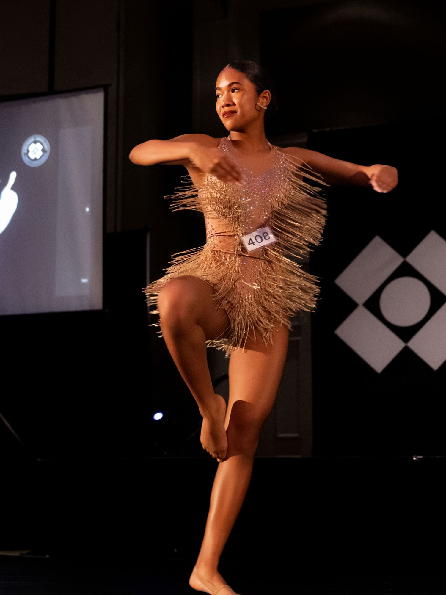 A dancer performs in a dark theater wearing a grass skirt-like costume with dramatic lighting and expressive movement.