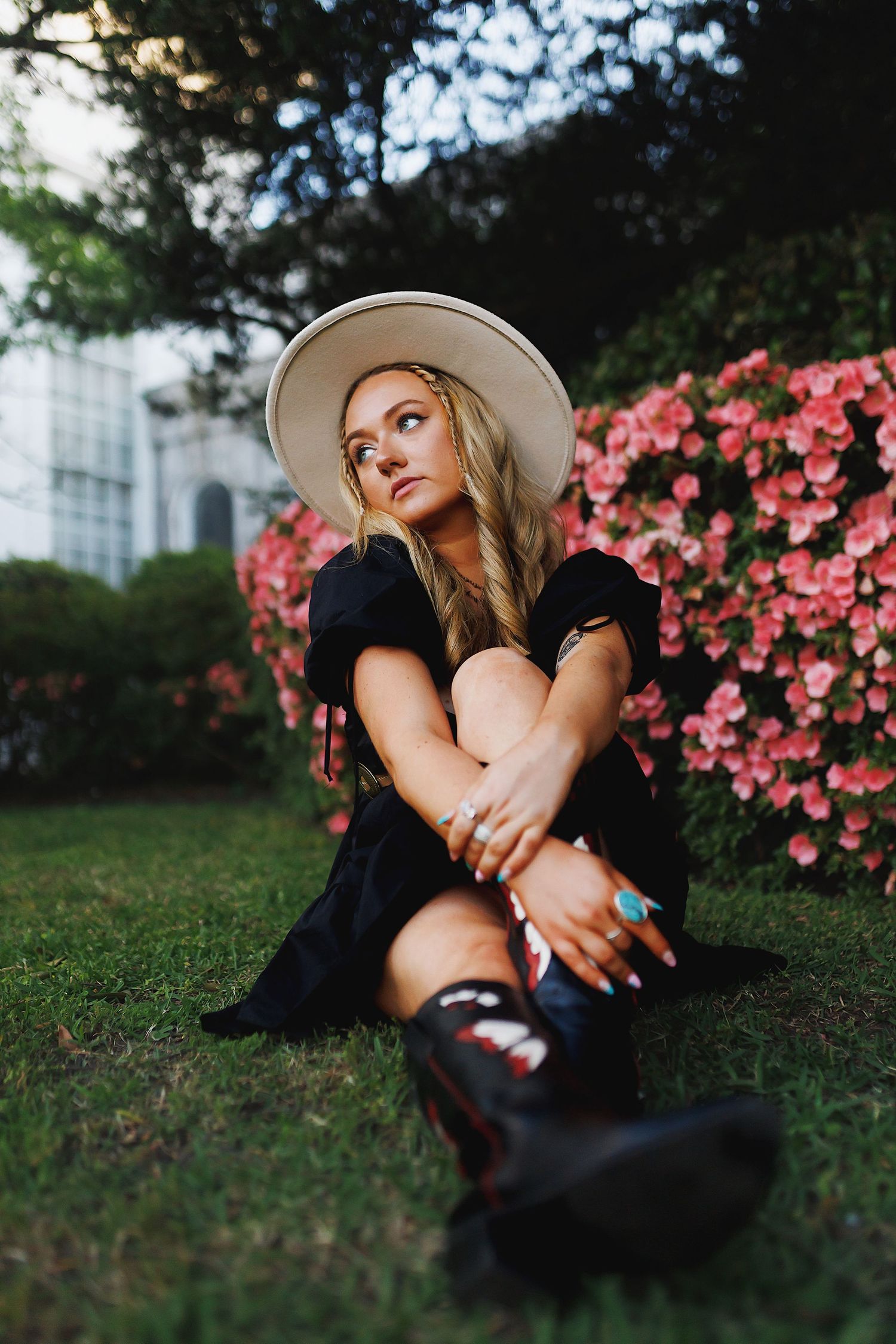 A dreamy outdoor portrait in a black dress and hat sitting by pink azalea bushes in a garden setting.