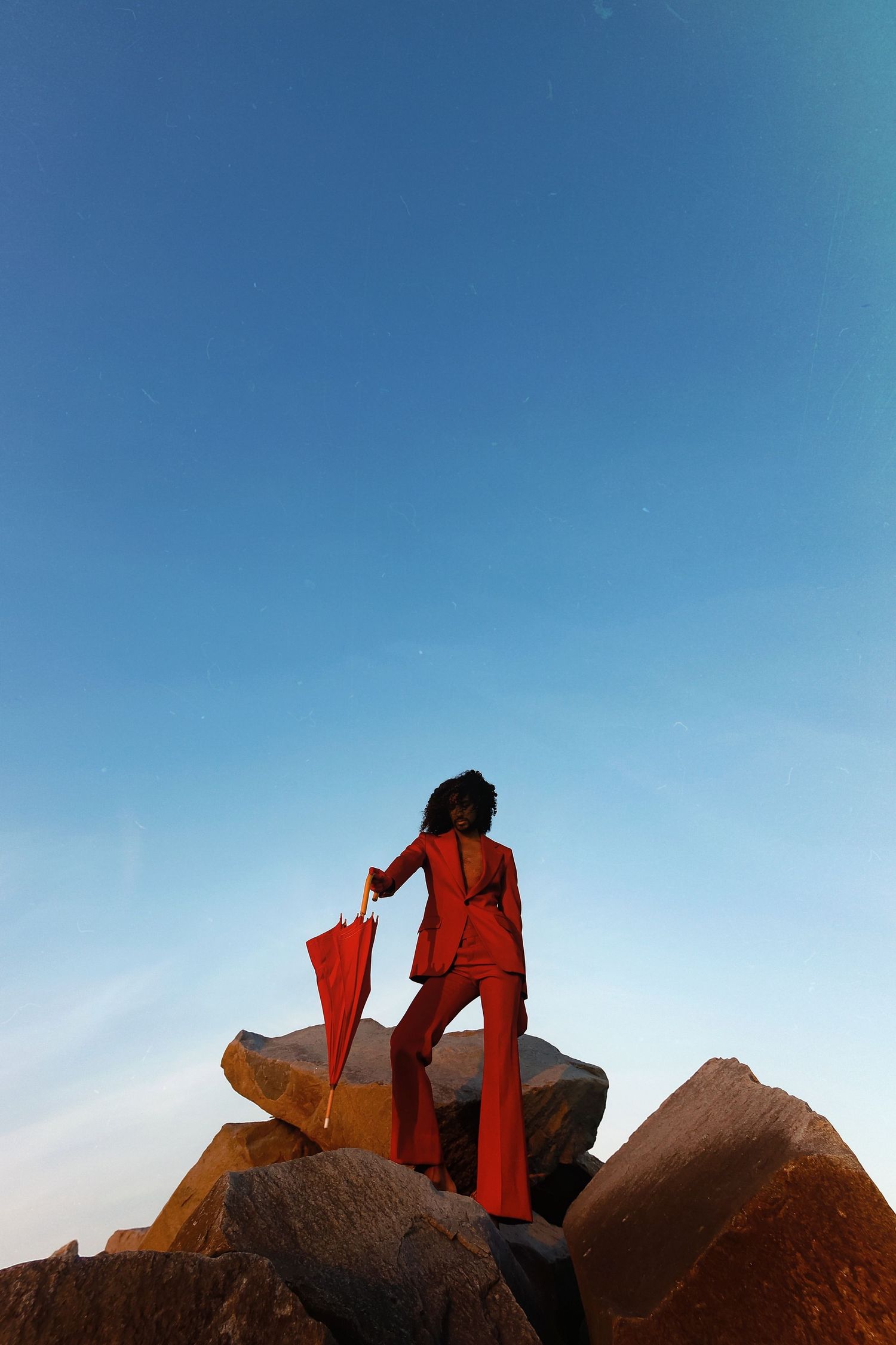A silhouetted figure in red sits perched atop a rocky outcrop against a bright blue sky.