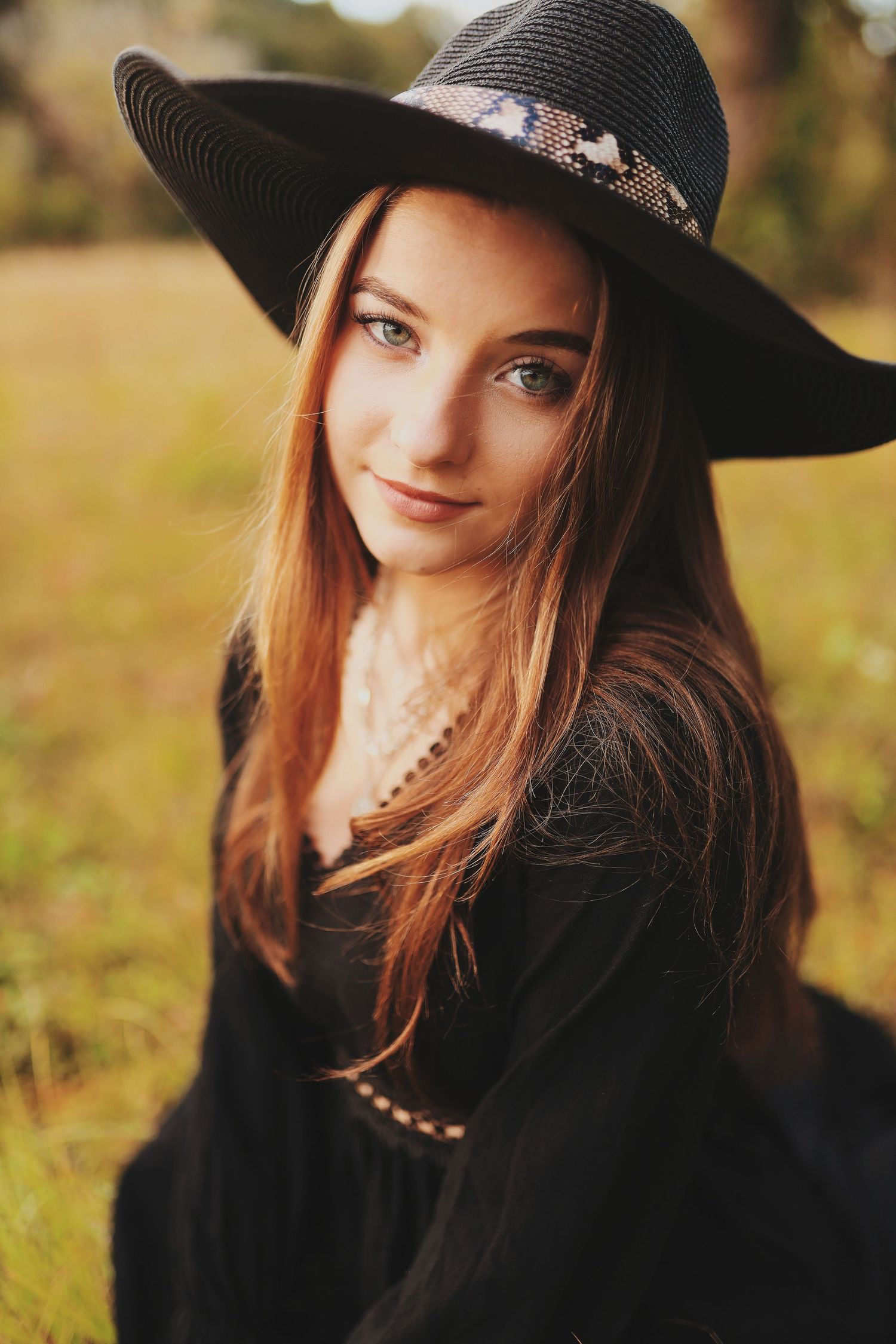 A person wearing a black wide-brimmed hat and dark clothing poses outdoors in autumn lighting.